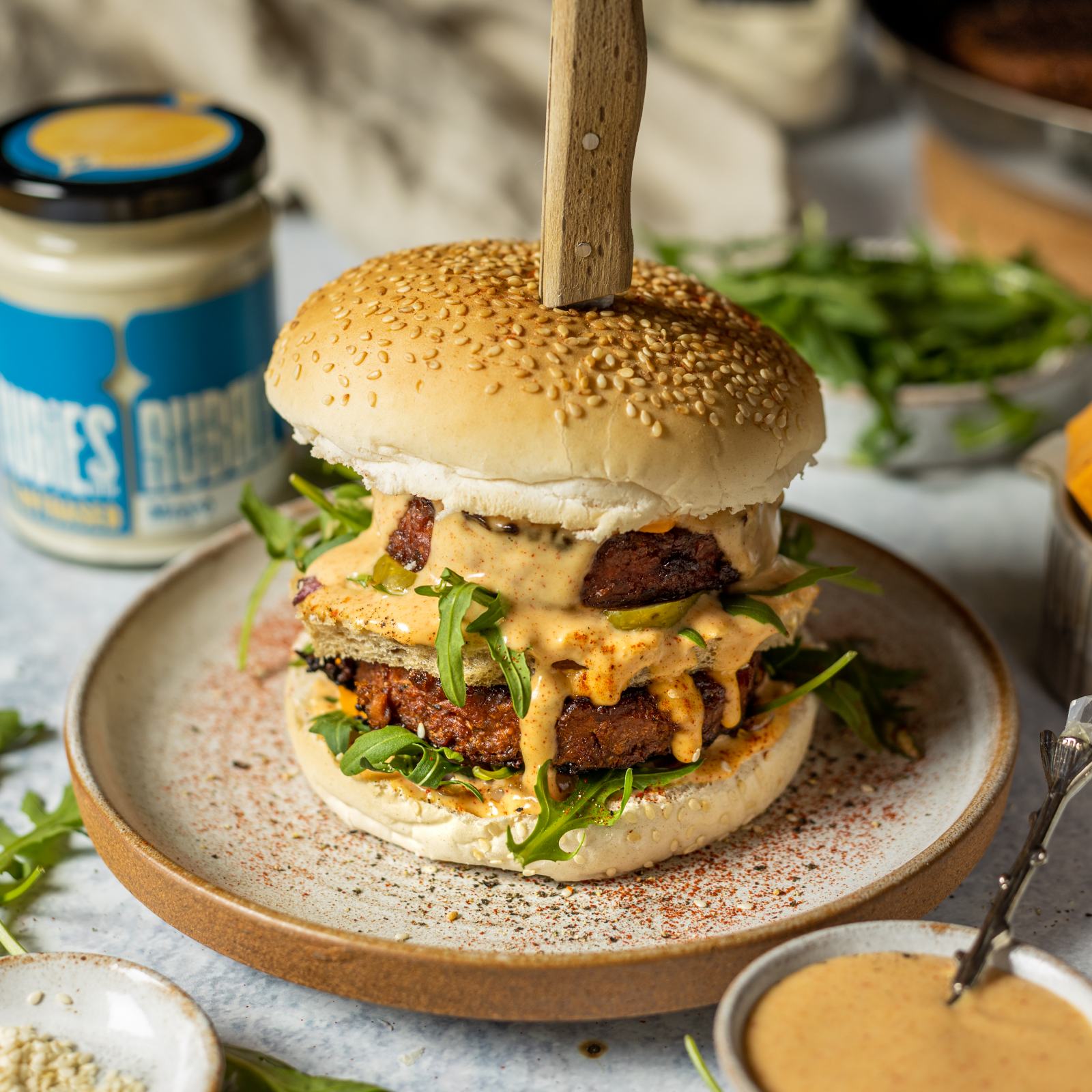 Vegan Mcdonalds Burger by a vegan big mac recipe in a seeded burger bun and 2 vegan burger patties on a white plate, surrounded by vegan burger ingredients