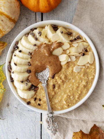 Vegan Pumpkin Spice Porridge photographed from above in a bowl, surrounded by little pumpkins and leaves, banana, nut butter and a spoon placed on top