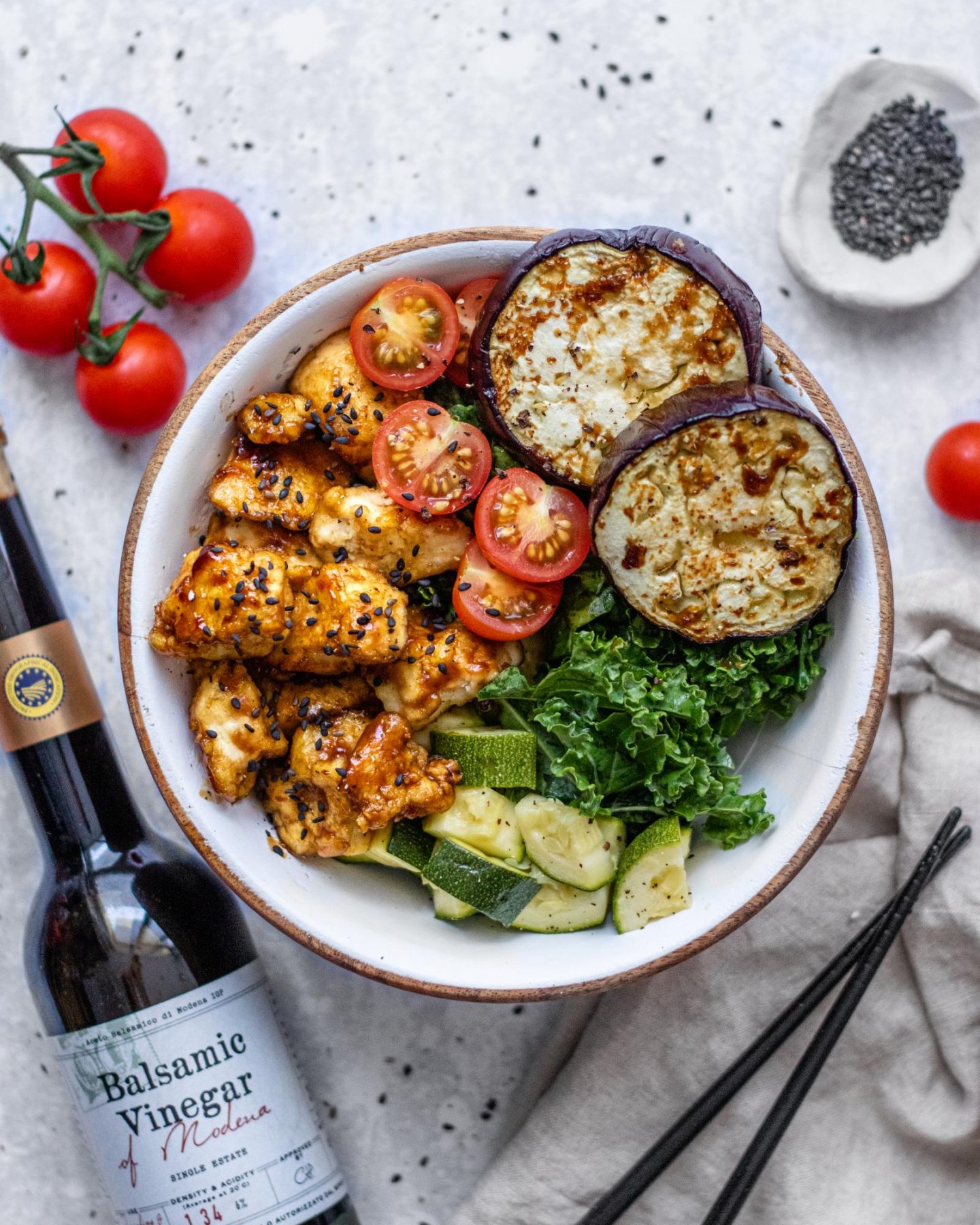 There is a bowl of roasted vegetables on a white table next to a bottle of balsamic vinegar. On top of the bowl we can see sticky balsamic tofu, tomatoes and roasted eggplant.