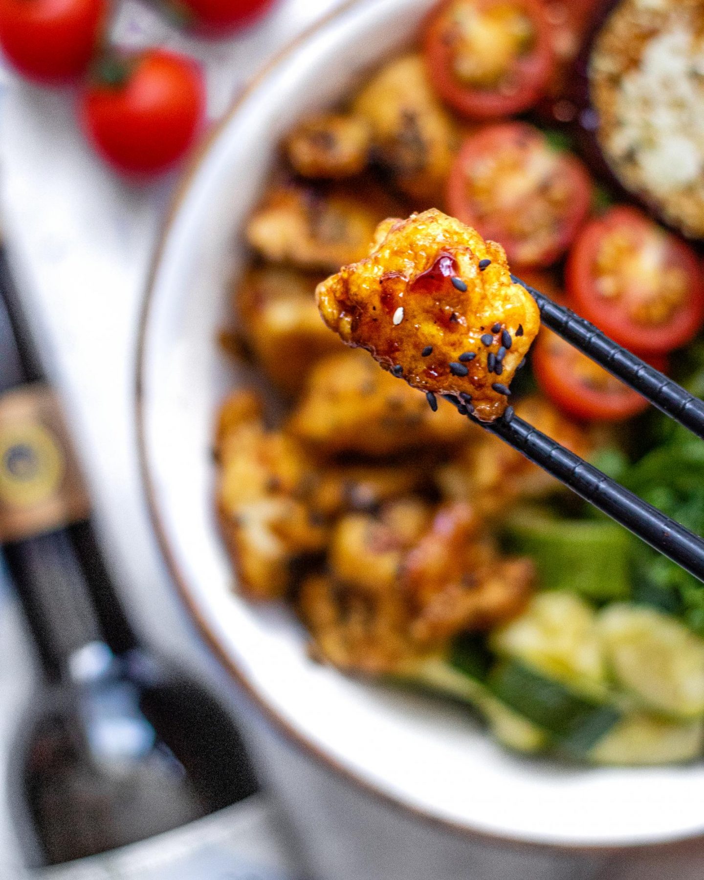 Two black chopsticks holding a piece of sticky balsamic tofu up close over a bowl of roasted vegetables.