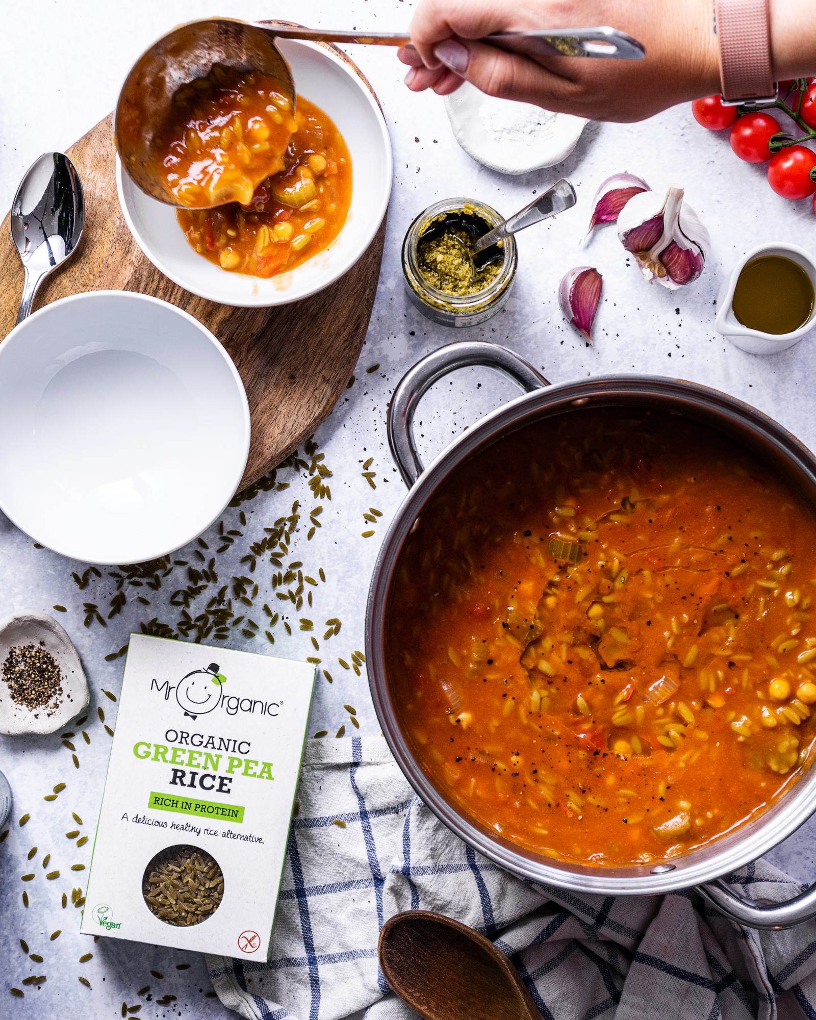 A large saucepan filled with orzo soup is sitting on a table and a hand is serving the soup into a bowl using a soup ladle.