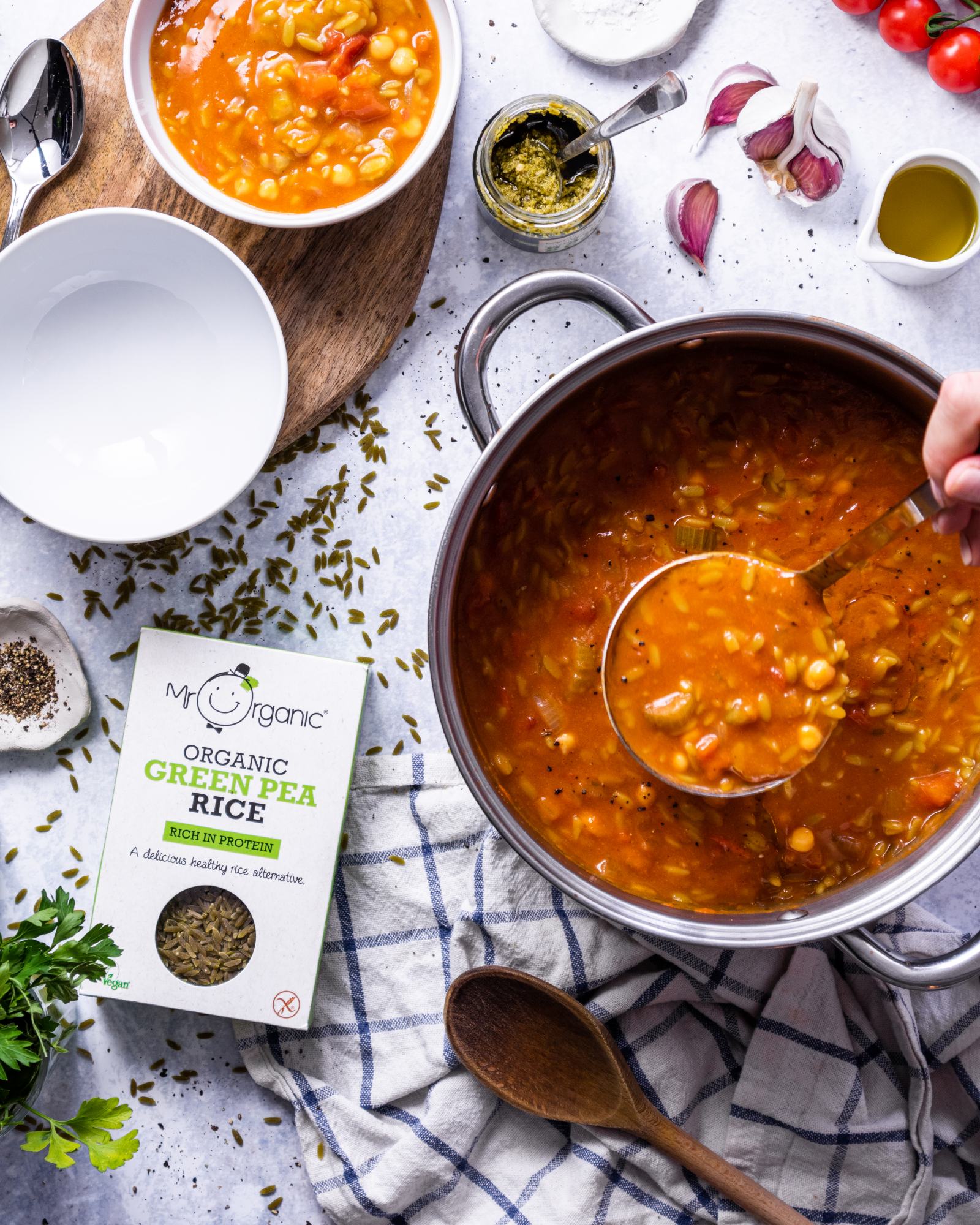 A large saucepan is filled with vegan orzo soup and a hand holding a soup ladle is lifting up a portion of the soup.