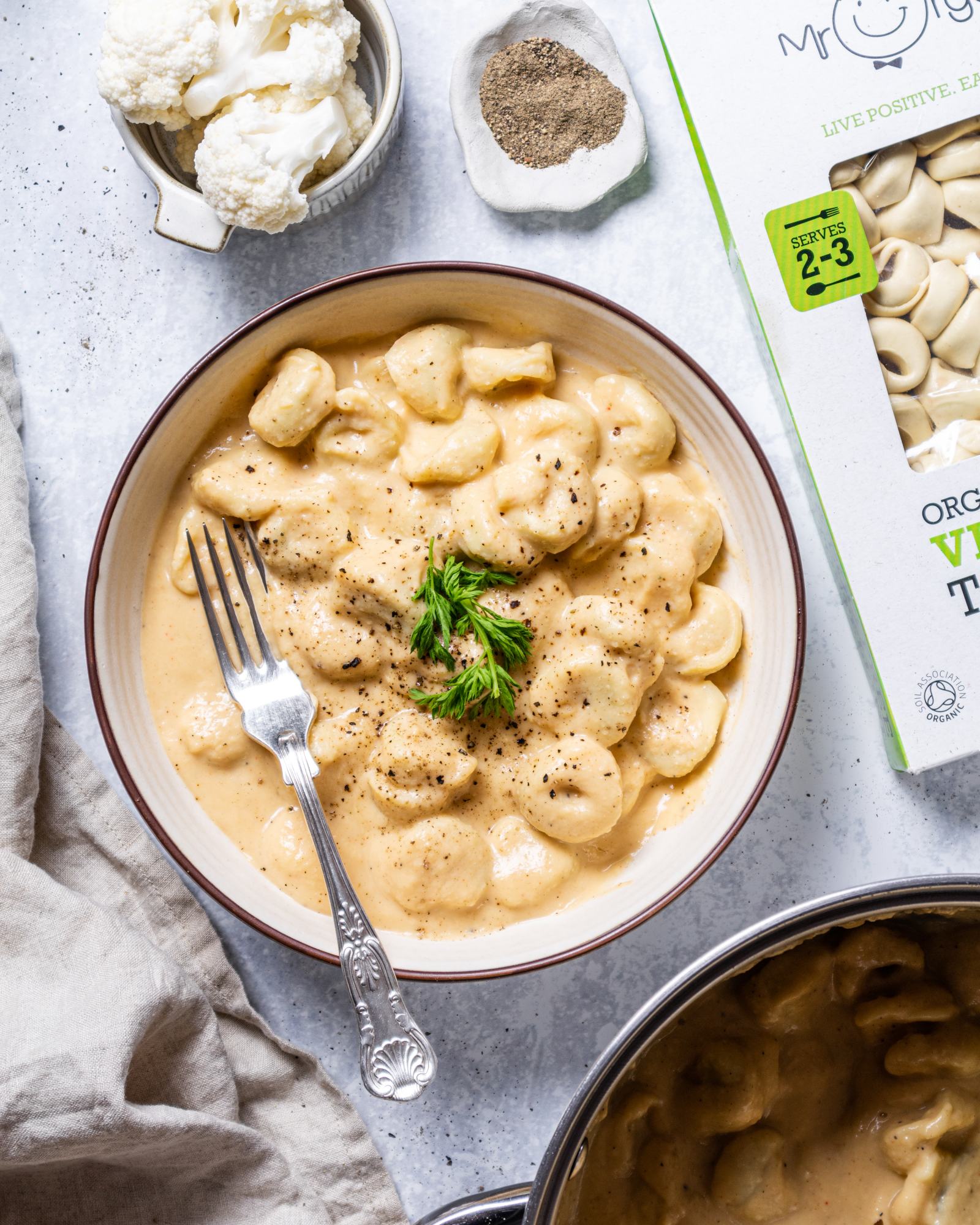 a portion of creamy vegan cauliflower alfredo on tortellini in a white serving bowl with herbs and black pepper on top and a fork on the side. The plate sits on a white table next to ingredients.