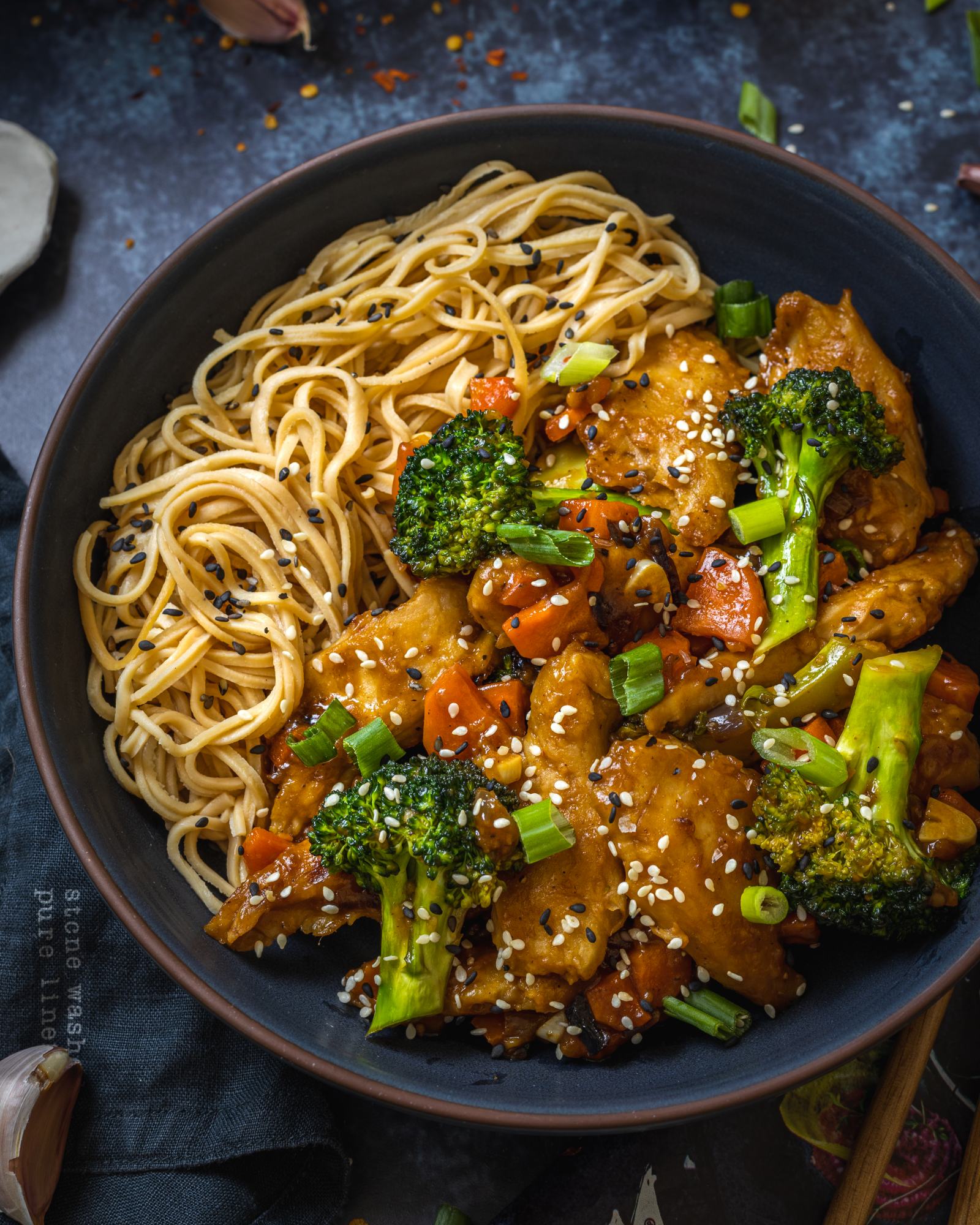 a black bowl on a black table filled with vegan teriyaki noodles and vegetables