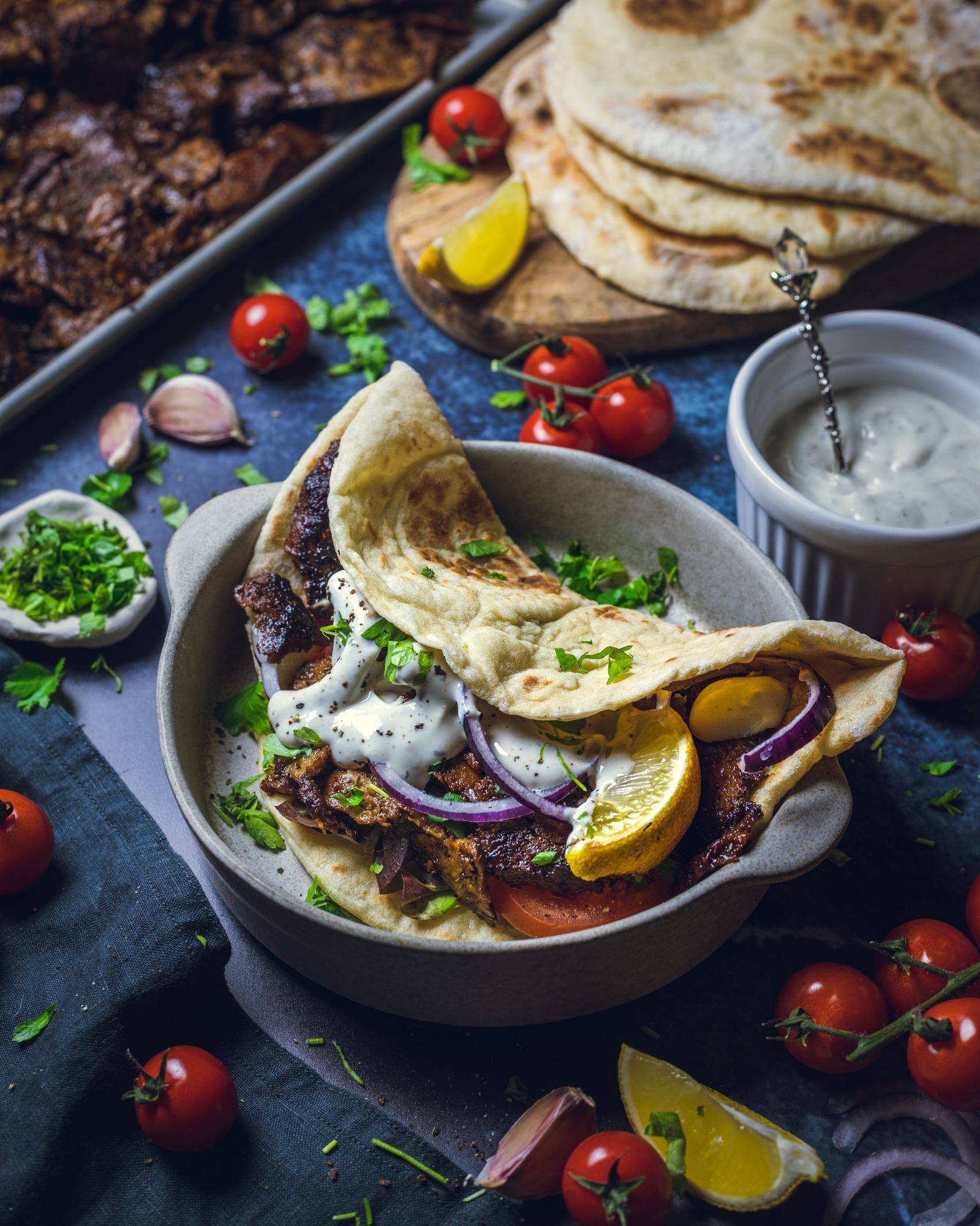 A homemade vegan Doner kebab with Seitan kebab meat, fresh tomatoes, lettuce and red onion is placed in a pitta on a dark table surrounded by ingredients. 