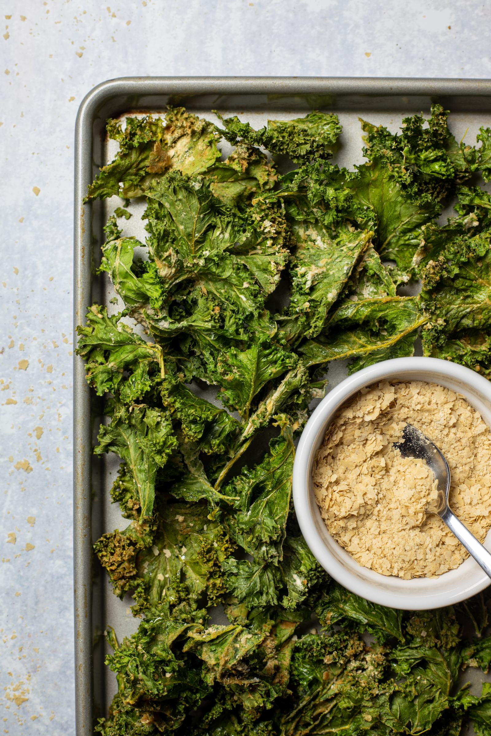 Healthy Kale Chips made in an air fryer are sitting on a serving board next to a bowl of cheesy nutritional yeast.