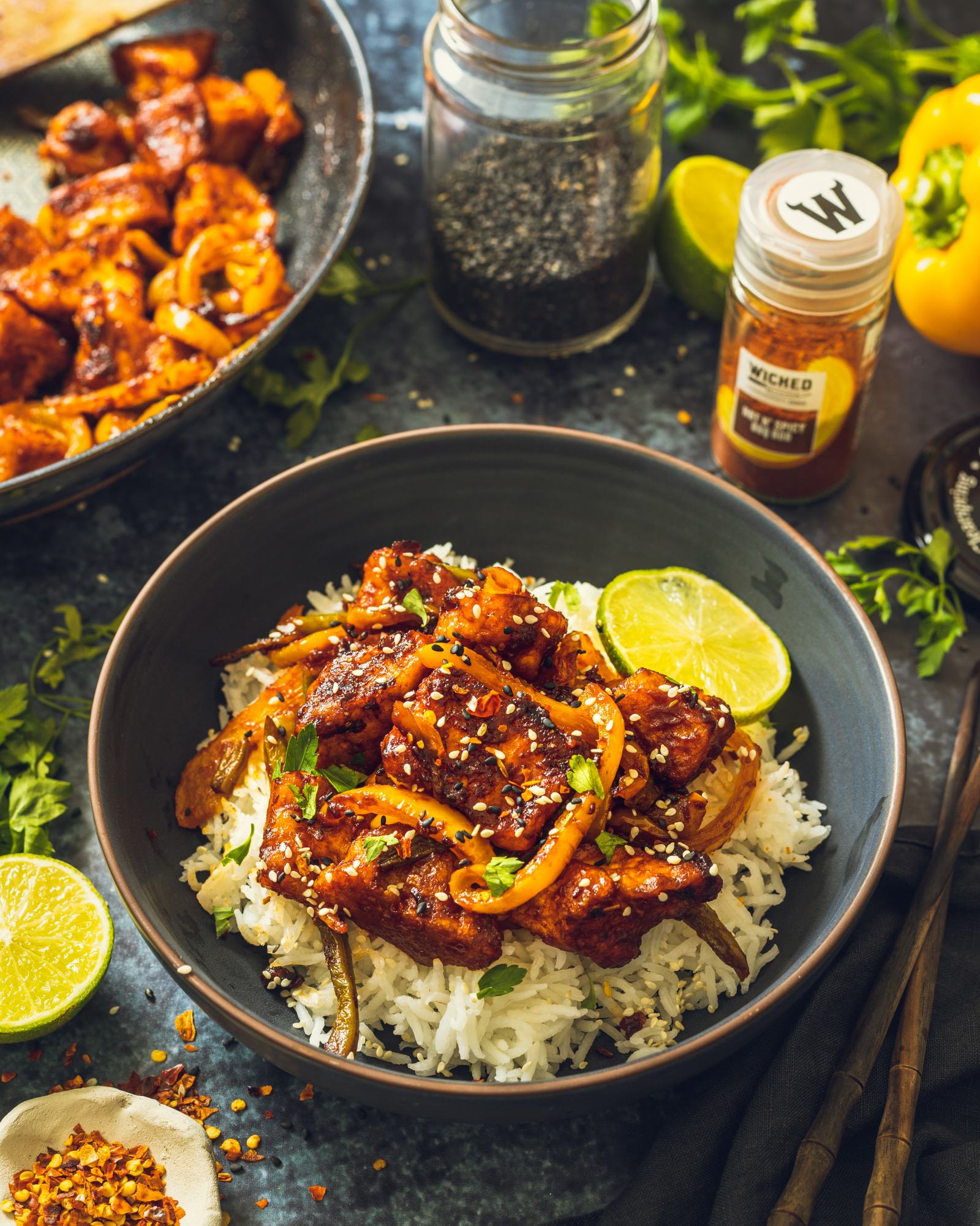 A bowl filled with fluffy rice and topped with vegan BBQ tofu is sitting on a dark table with ingredients surrounding the bowl.