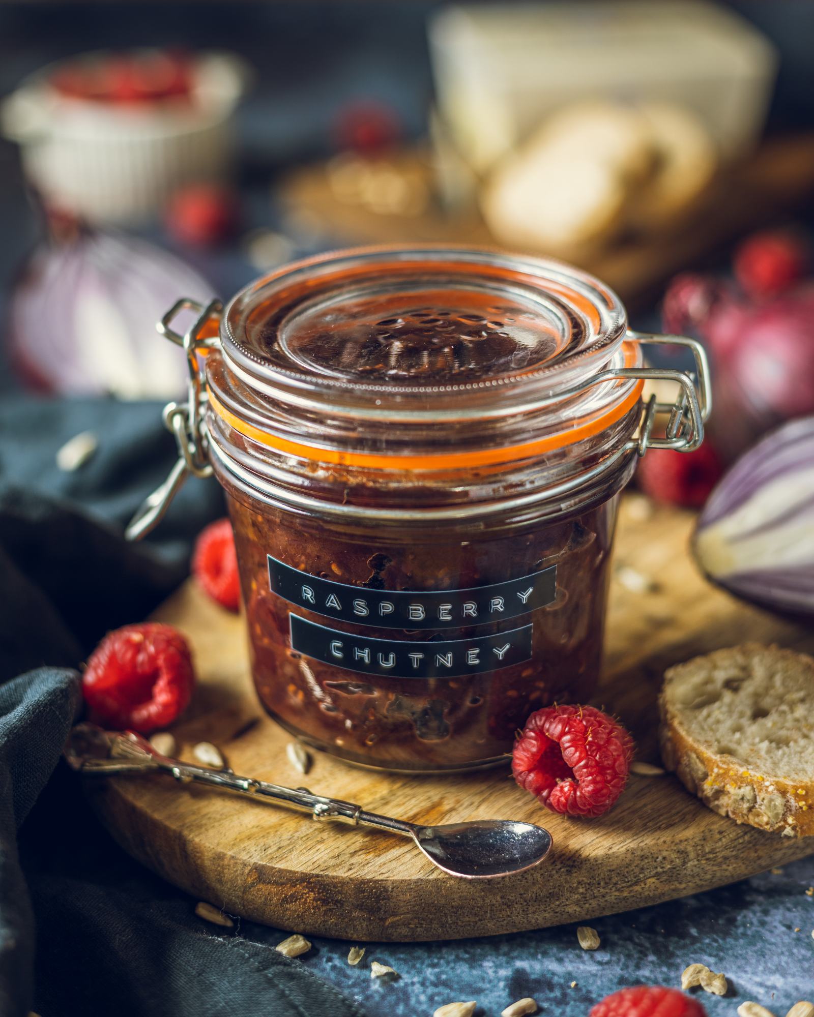 The full jar of raspberry red onion chutney on a wooden serving board surrounded by ingredients such as raspberries red onions.