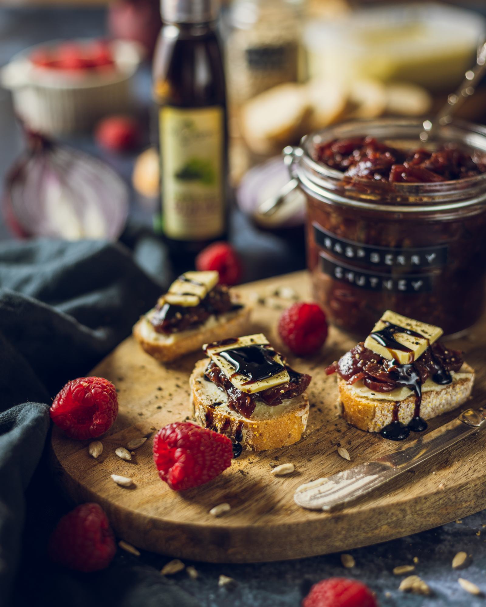 Small bites of bread crackers topped with raspberry red onion chutney small slices of vegan cheese and a balsamic drizzle on a serving board surrounded by ingredients and with the chutney jar in the background.