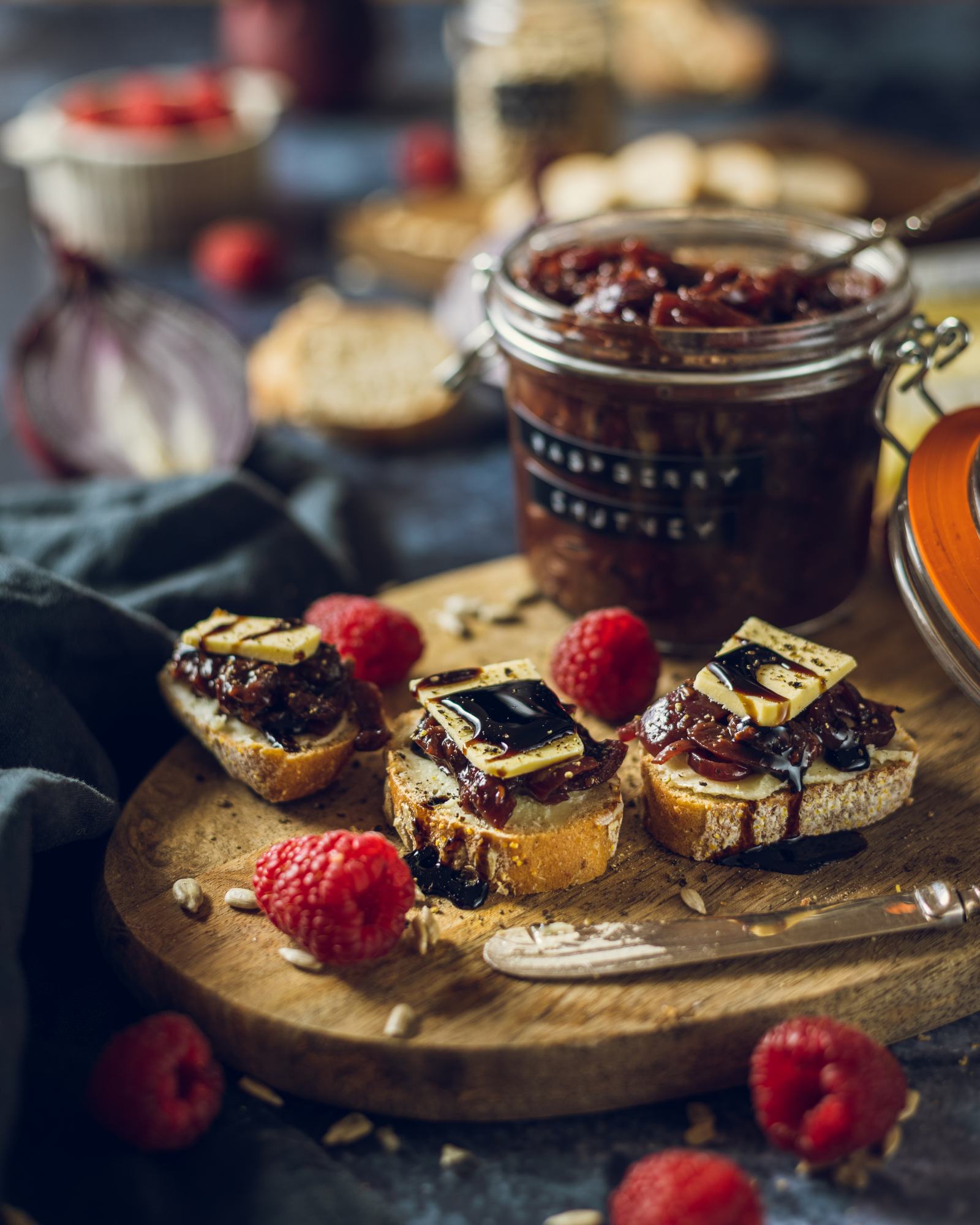 Raspberry and red onion chutney on small slices of bread alongside vegan cheese with the full jar of raspberry chutney visible in the background.