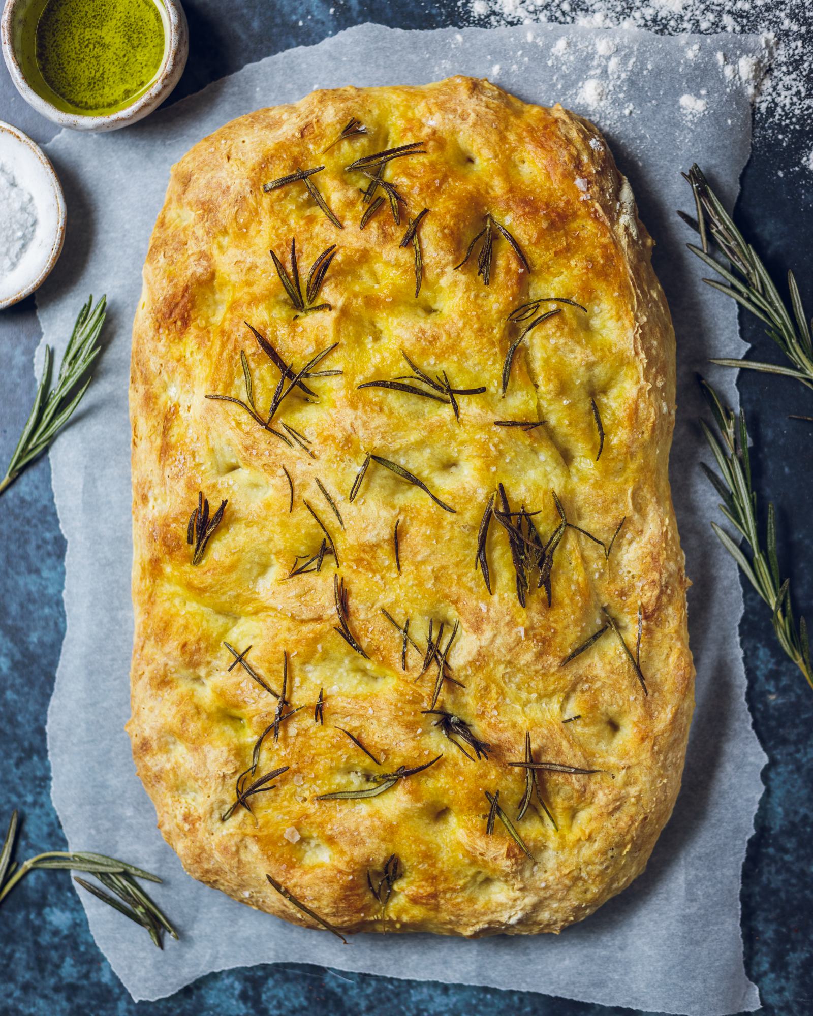 Freshly baked rosemary focaccia from above on a baking parchment with fresh rosemary, salt and olive oil next to it.