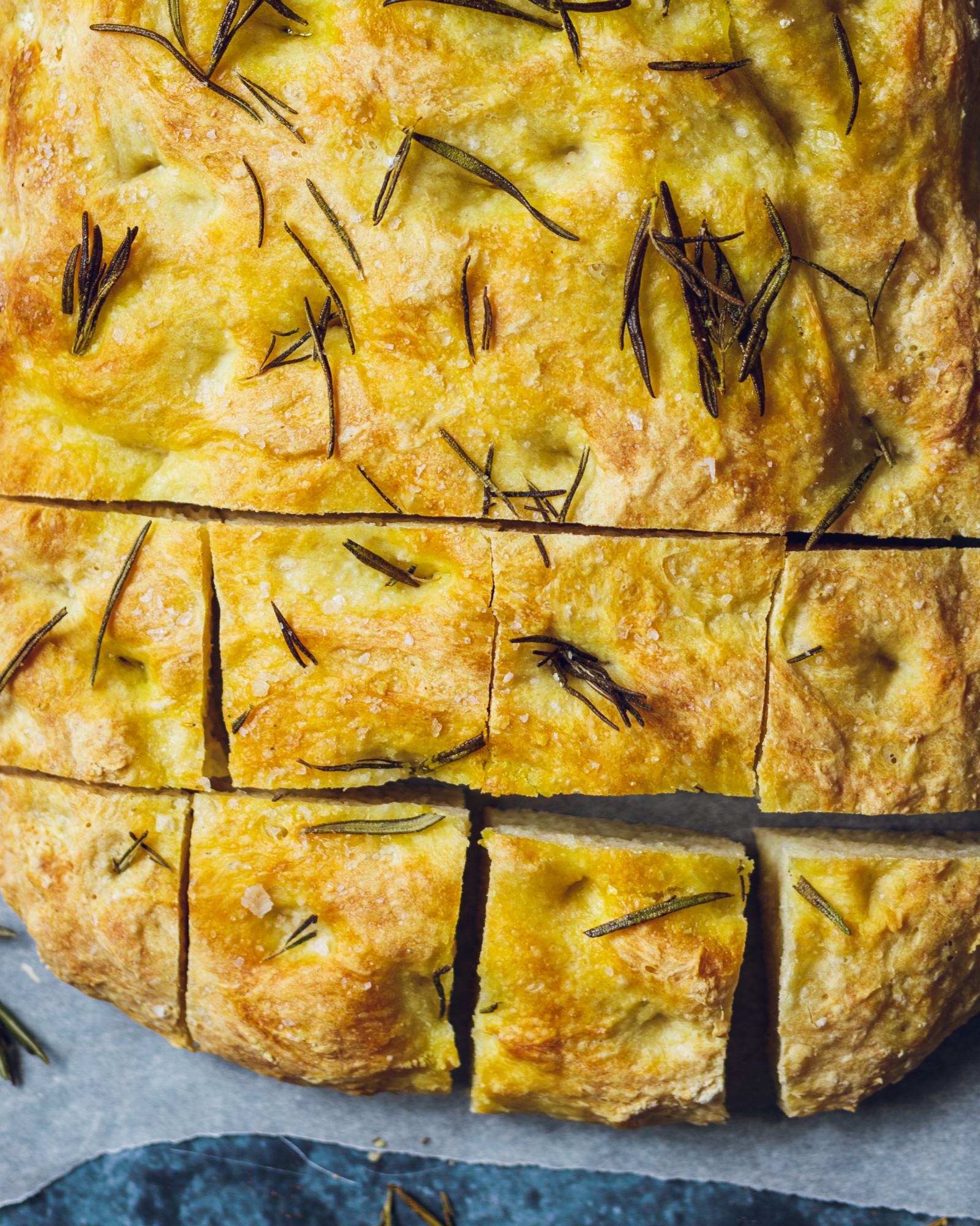 A loaf of vegan rosemary focaccia on baking parchment with the bottom half sliced into cubes.