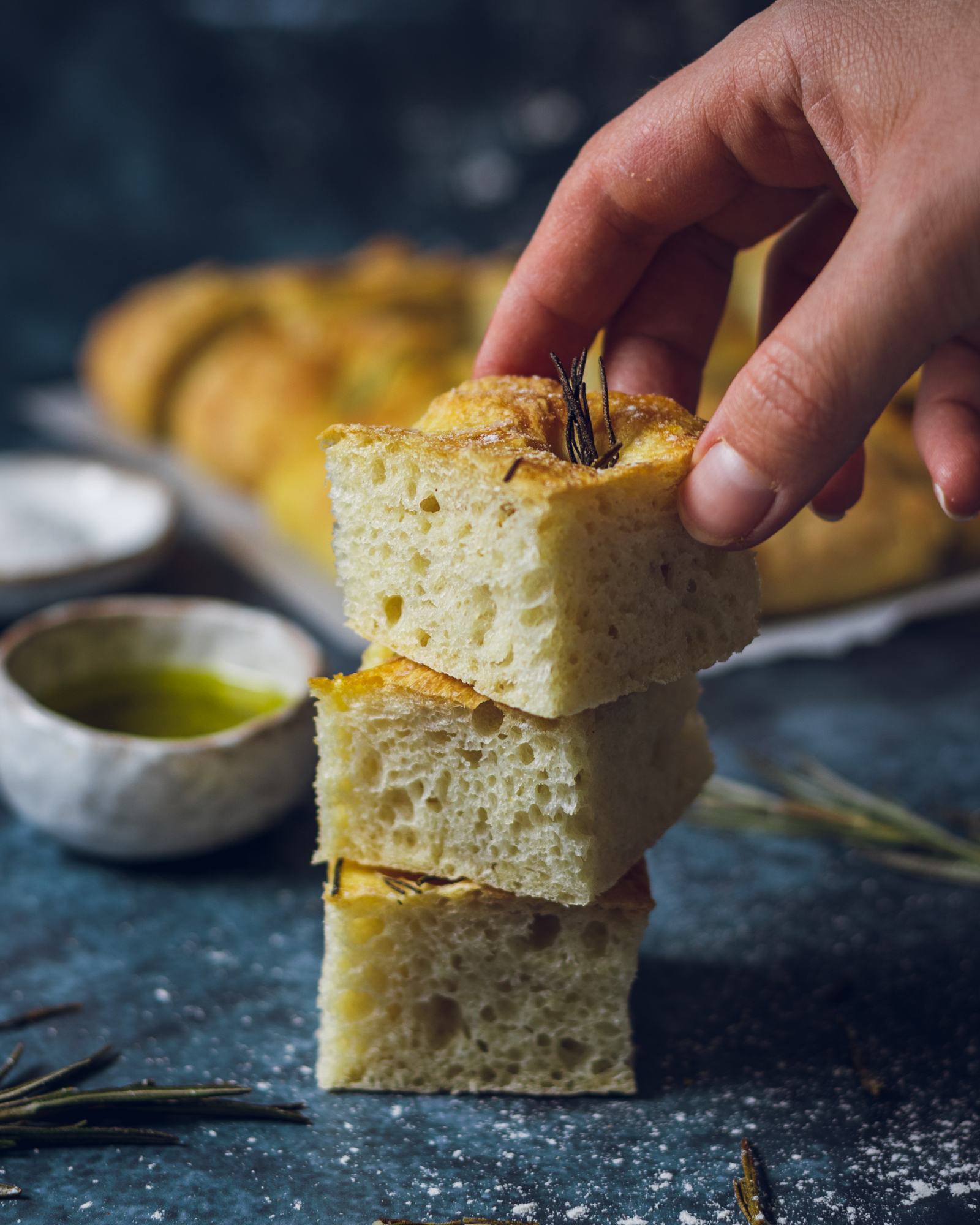 A hand stacking 3 pieces of focaccia on top of each other on a dark table. 