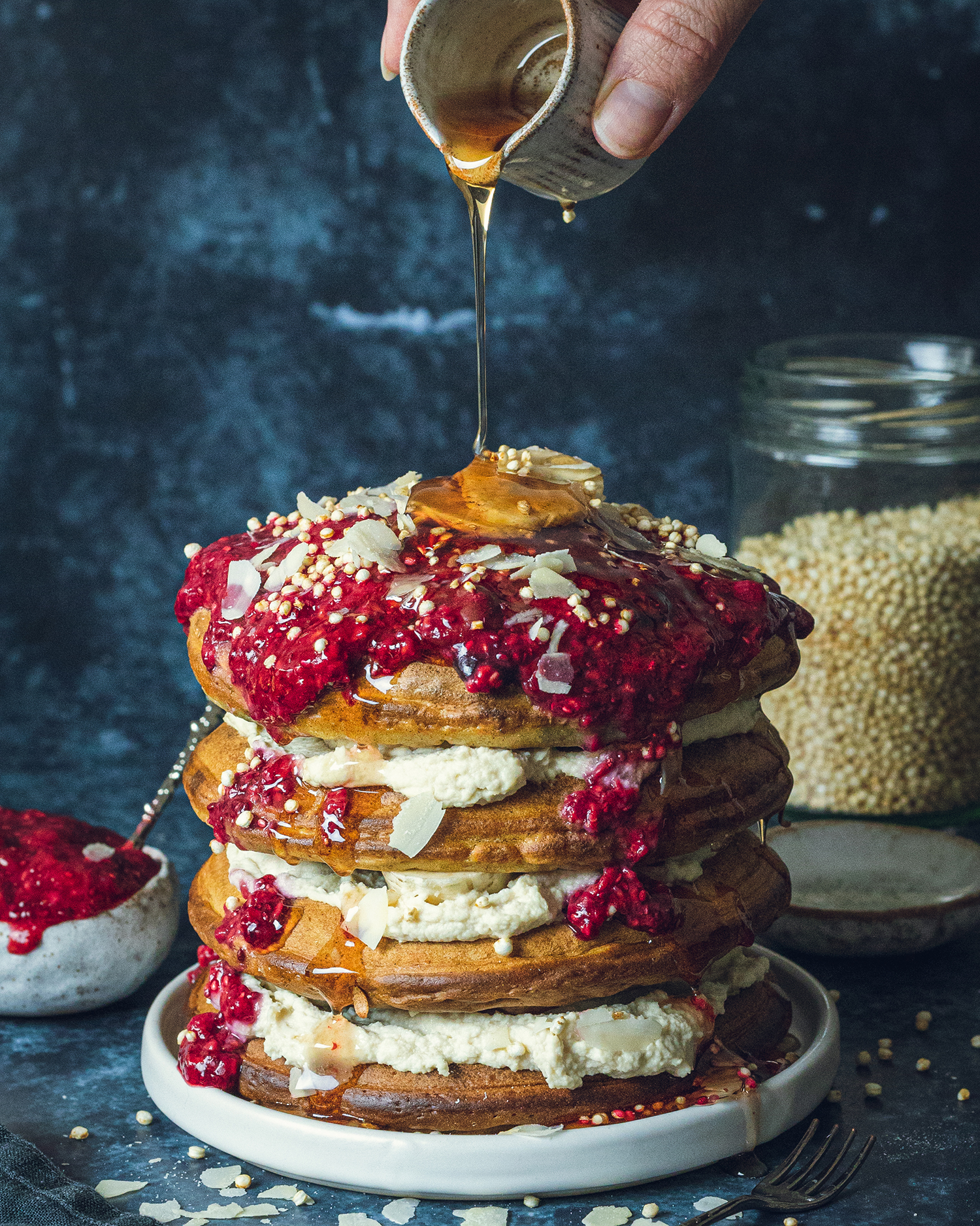 a stack of vegan buttermilk pancakes with yogurt and chia jam and a hand drizzling maple syrup from a small jug over the top.