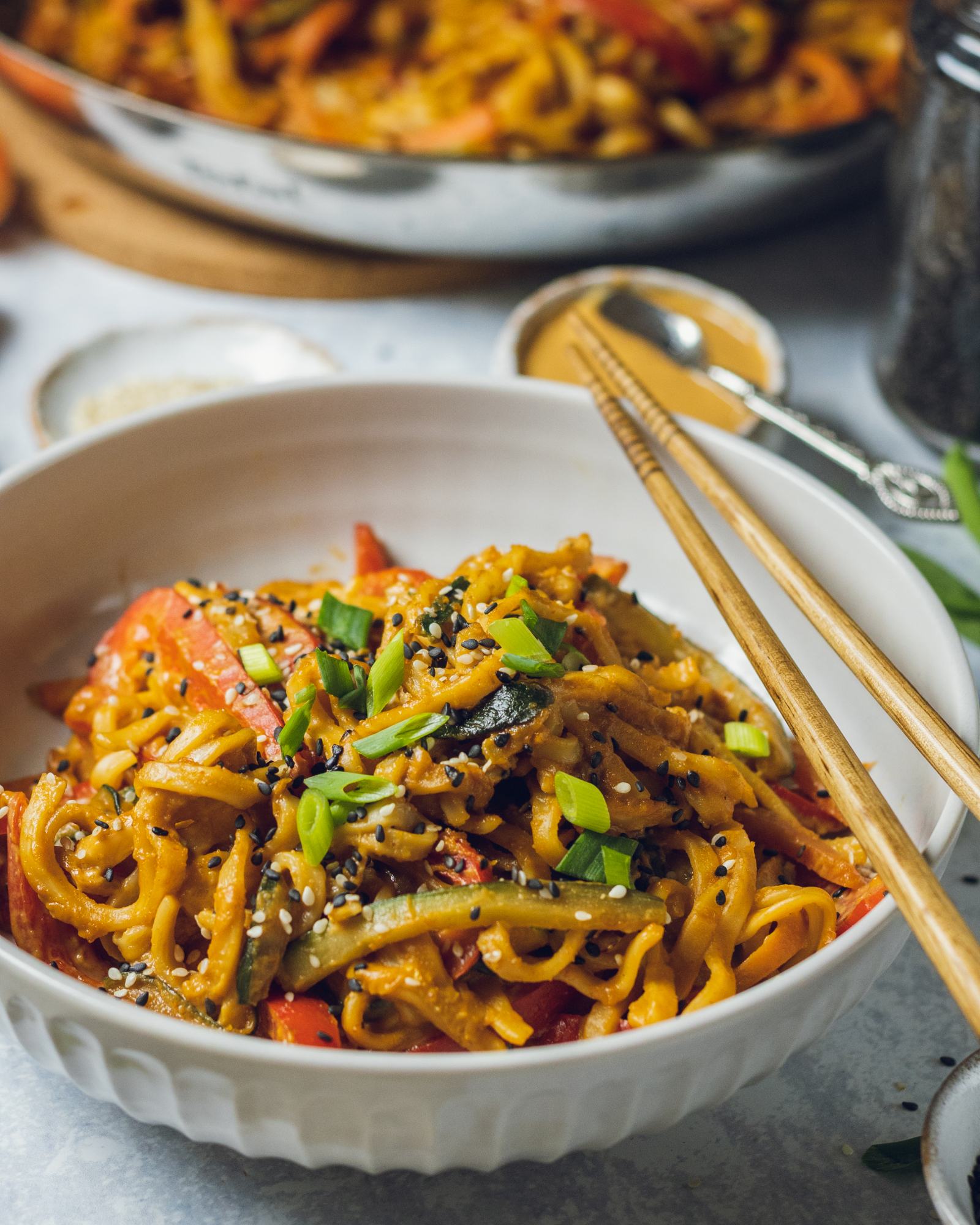 Creamy peanut noodles in a white serving bowl can be seen on the on the dinner table with chopsticks resting on the side.