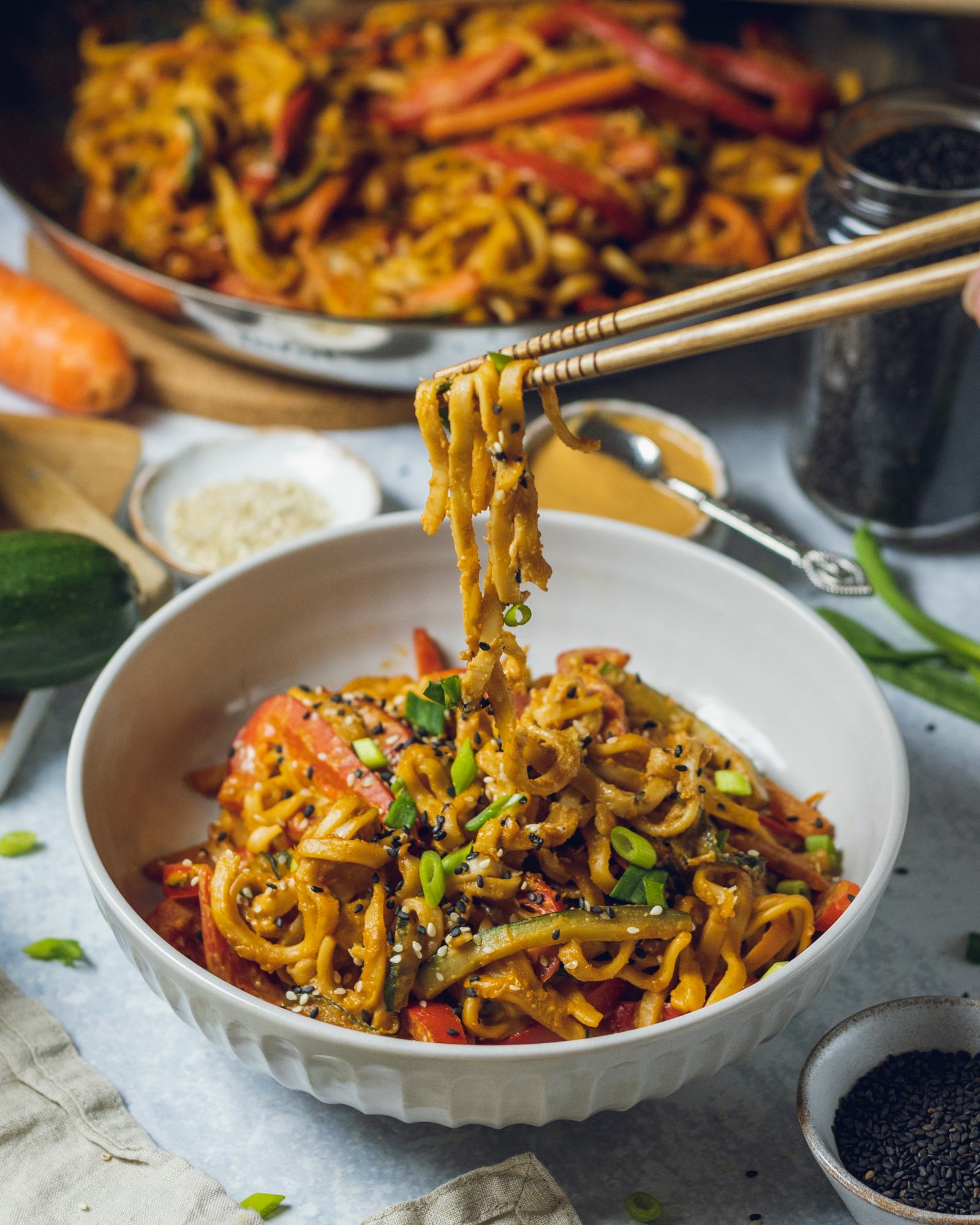 A white bowl filled with udon noodles in a creamy peanut sauce alongside peppers, zucchini and green onions can be seen on a dinner table surrounded by ingredients. Wooden chopsticks are picking up a portion of the noodles.