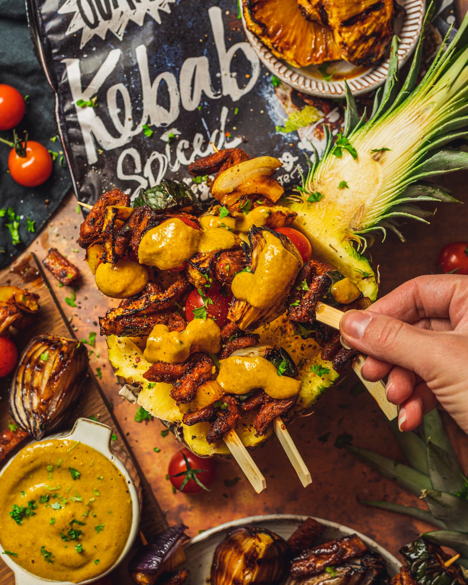 a hand reaching for skewers served on a hollow pineapple half with Pineapple Curry BBQ Sauce drizzled over the top