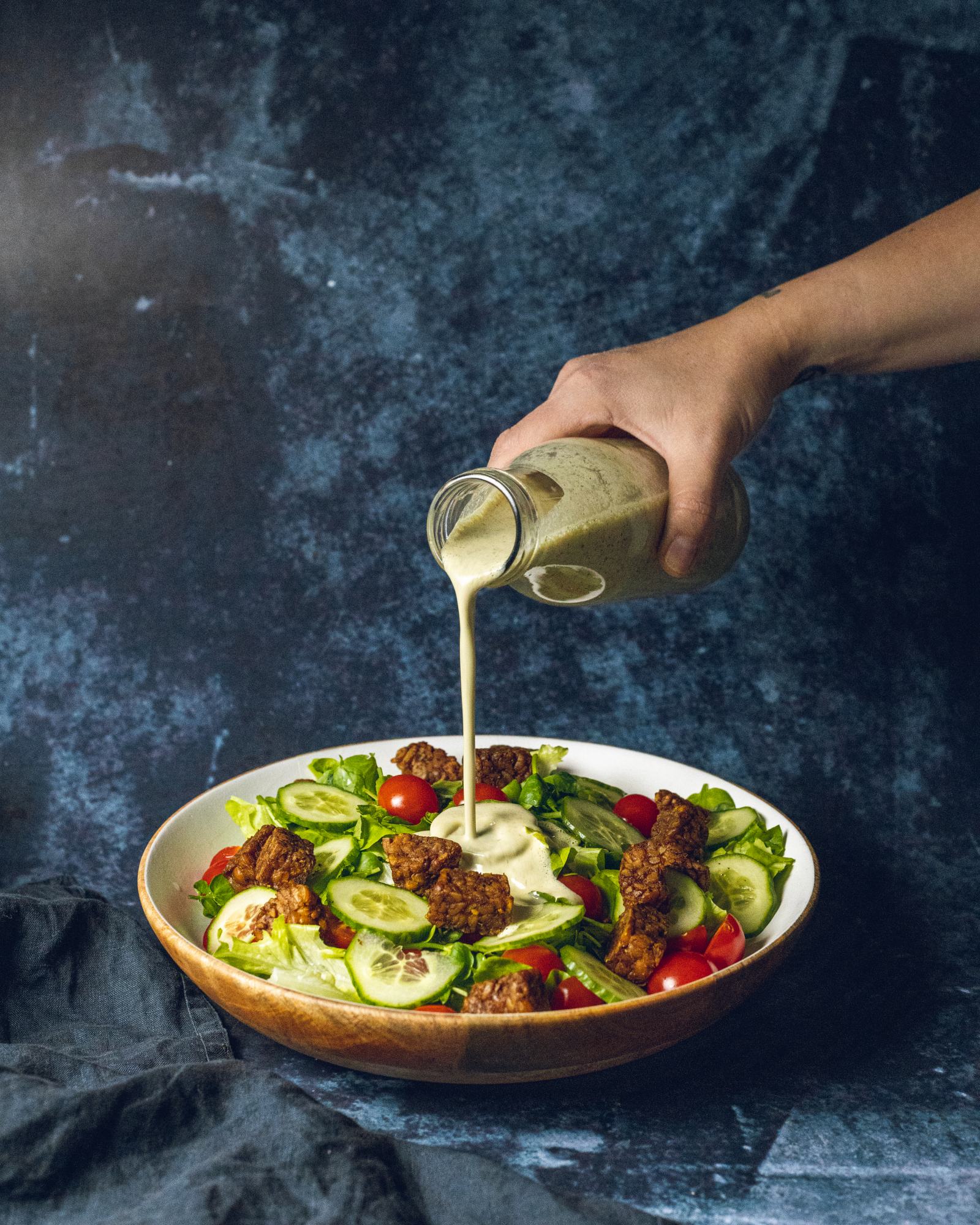 A hand is pouring vegan Caesar Salad dressing from a glass bottle onto a large salad bowl.