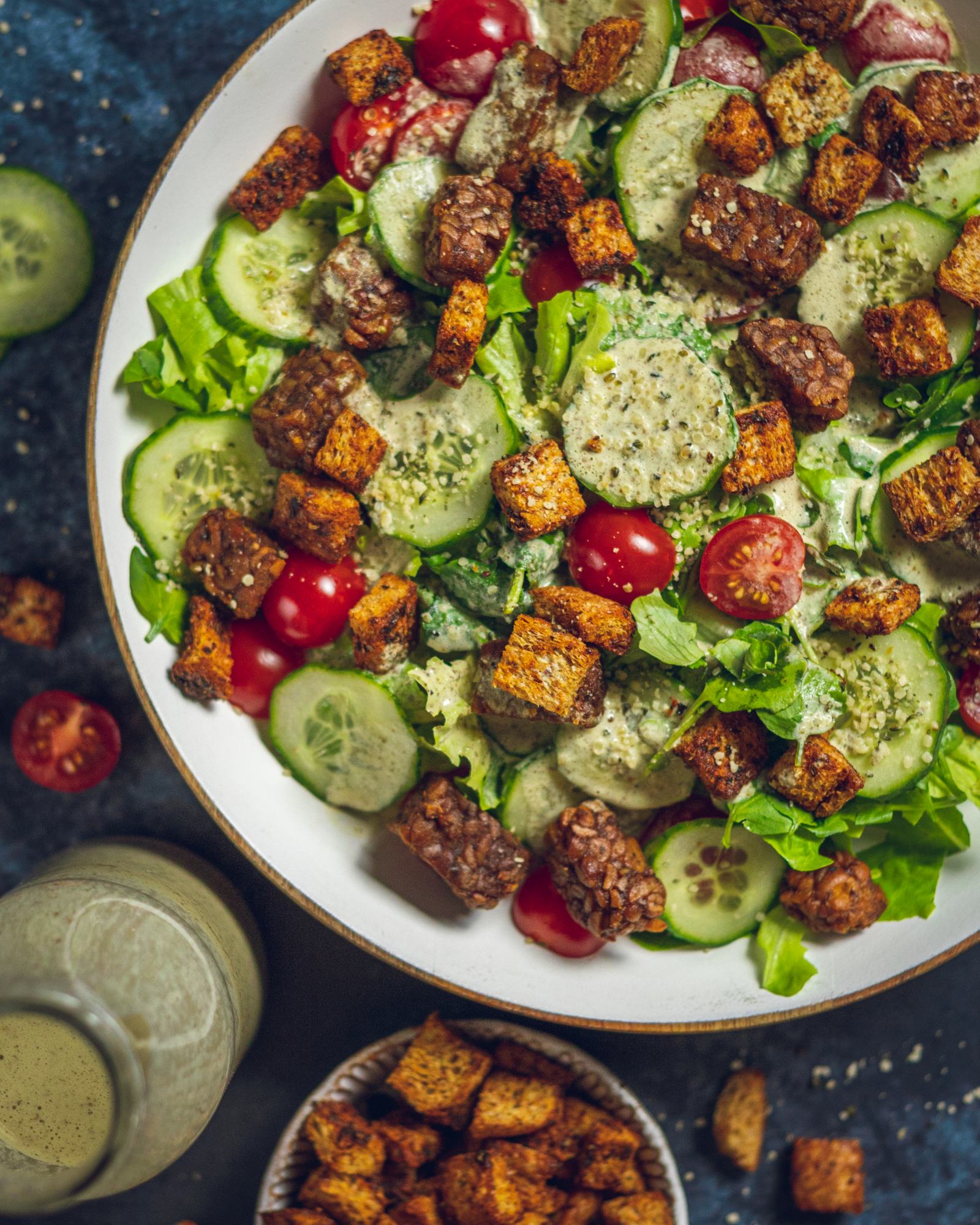 A large salad bowl is sitting on a dark blue table next to salad dressing and ingredients. There are crunchy croutons placed on top of the salad. 