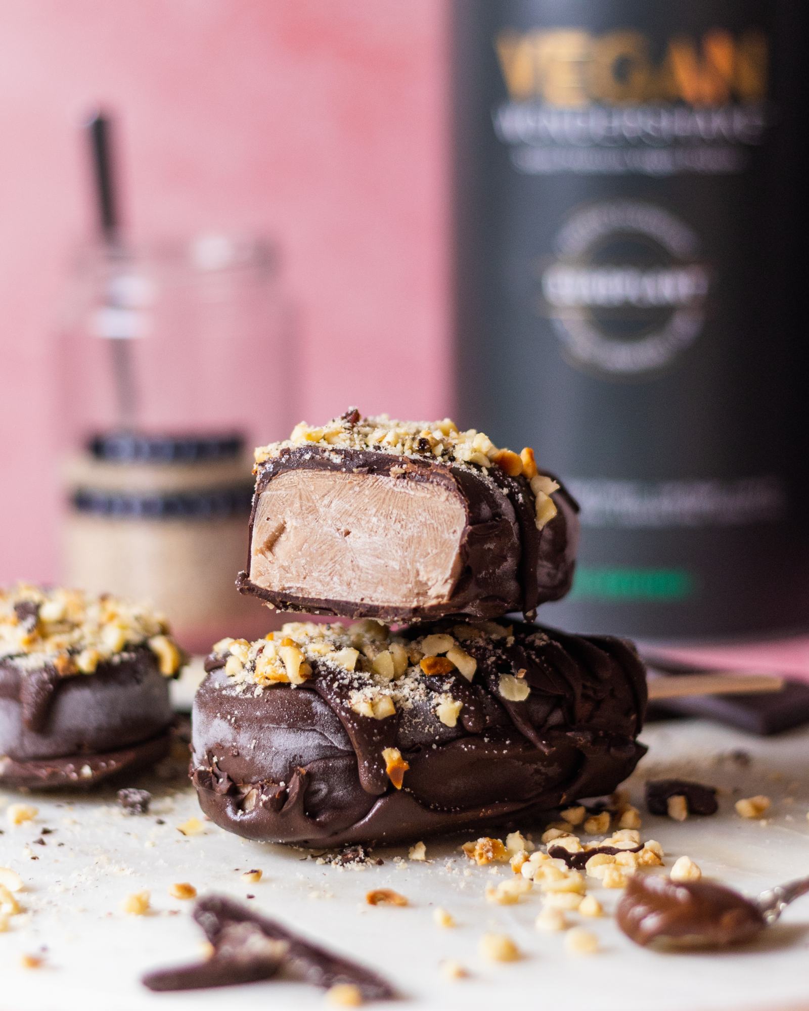 Vegan Chocolate Hazelnut Magnum Ice Cream, one cut open laying on top of another one, protein tub blurry in the background, on pink backdrop