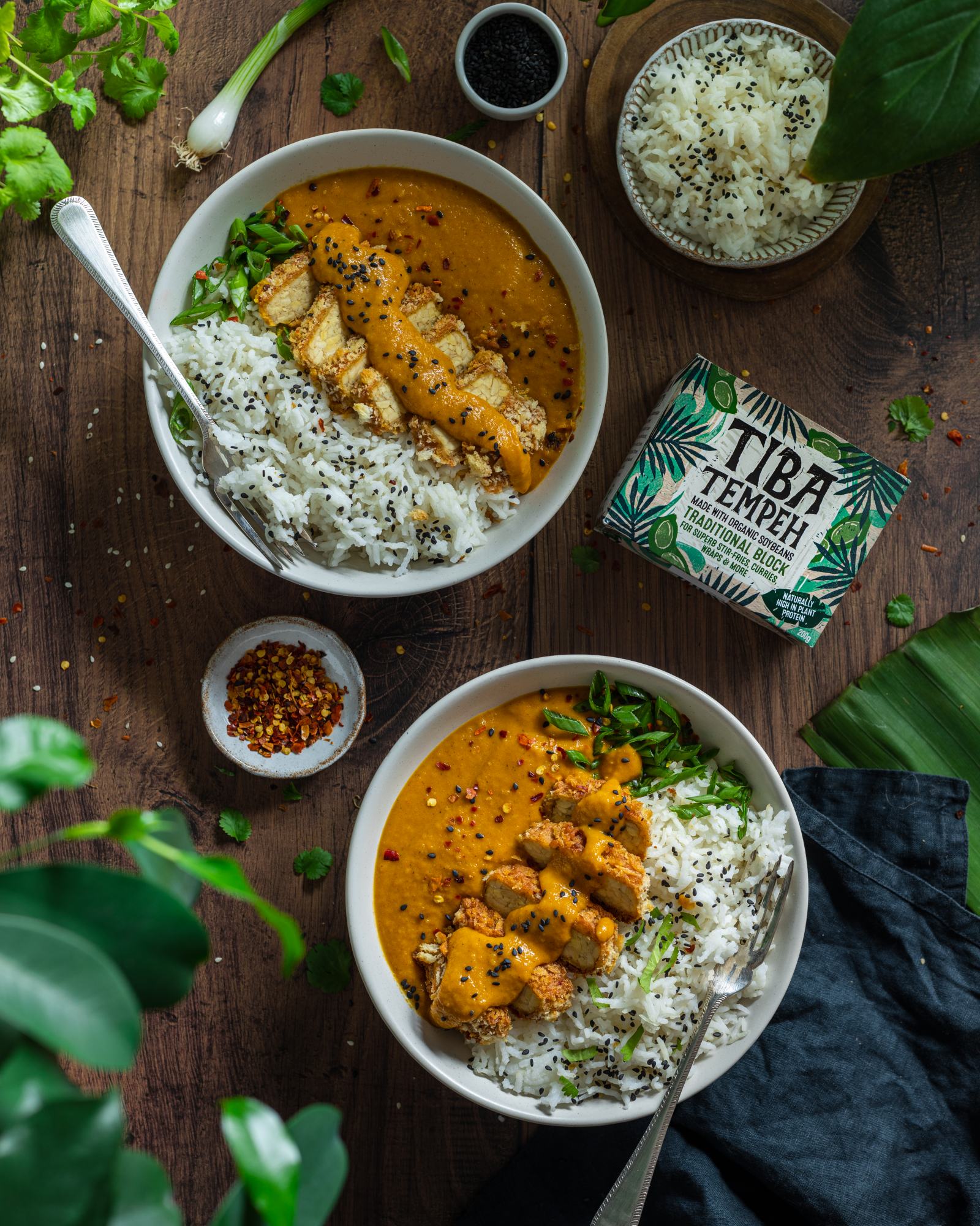 Vegan Katsu Curry with Tempeh - two bowls on a dark backdrop, rice and katsu sauce on each bowl with a fork, sliced crunchy panko coated tempeh on top with a drizzle of sauce, tiba tempeh packaging placed besides the bowls