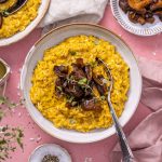 Vegan Pumpkin Risotto in a white bowl with mushrooms on top and a spoon on the side, photographed on a pink backdrop