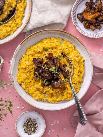 Vegan Pumpkin Risotto in a white bowl with mushrooms on top and a spoon on the side, photographed on a pink backdrop