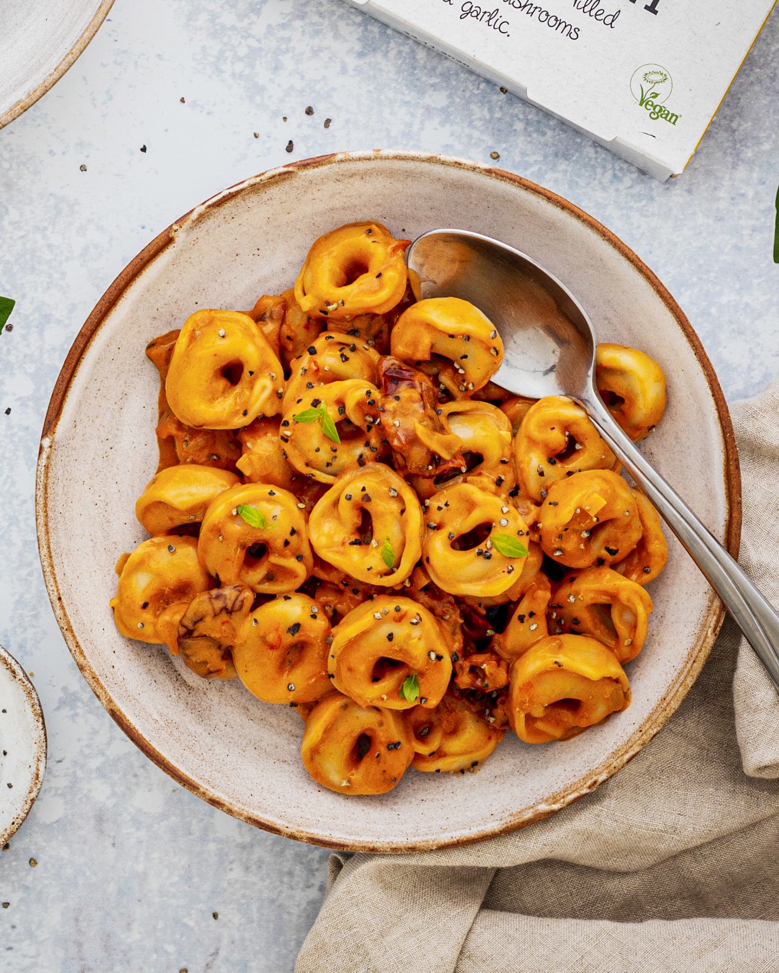 Creamy Sun-Dried Tomato Pasta - Vegan Tortellini on a plate with a spoon placed on the side and a napkin under the bowl, on a bright table top