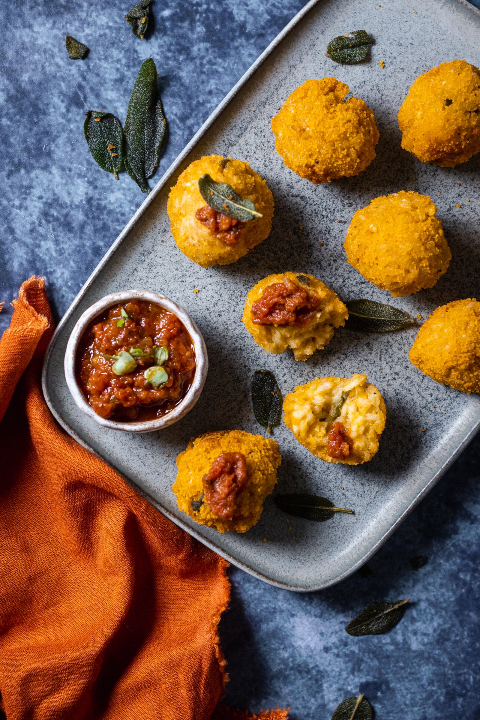 crunchy vegan pumpkin arancini on a serving platter with a dip on the side and crispy sage leaves on top