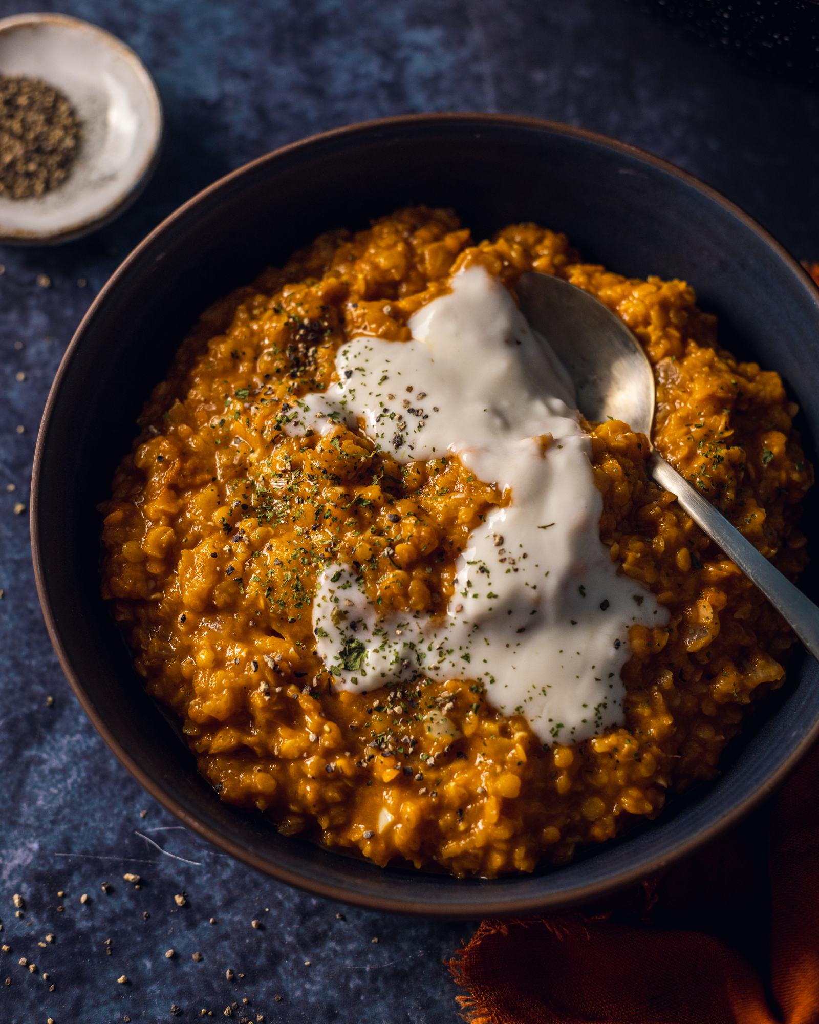 a plate full of Roasted Pumpkin Red Lentil Dhal placed on a dark blue background, with a spoon and coconut yoghurt on the top
