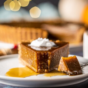 a slice of vegan pumpkin pie on a white plate with a fork having taken the tip off. the fork if laying next to the piece of pie and you can see the rest of the vegan pumpkin pie in the background