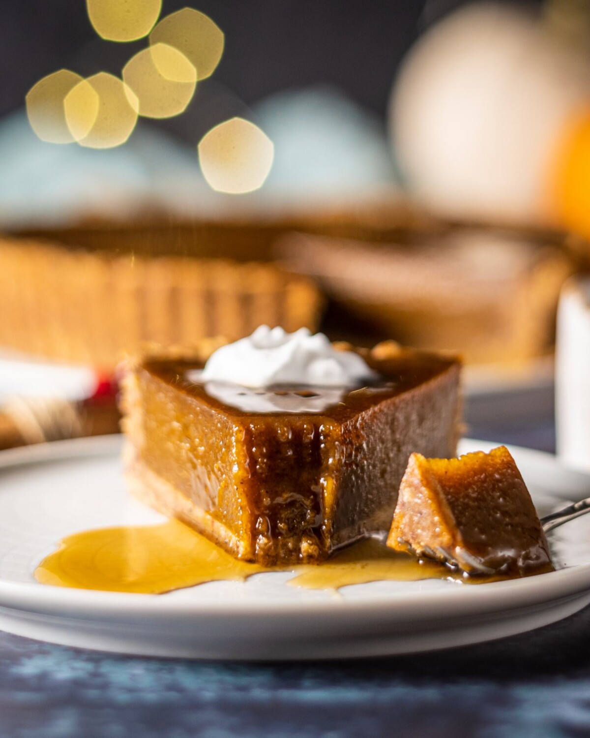 a slice of vegan pumpkin pie on a white plate with a fork having taken the tip off. the fork if laying next to the piece of pie and you can see the rest of the vegan pumpkin pie in the background