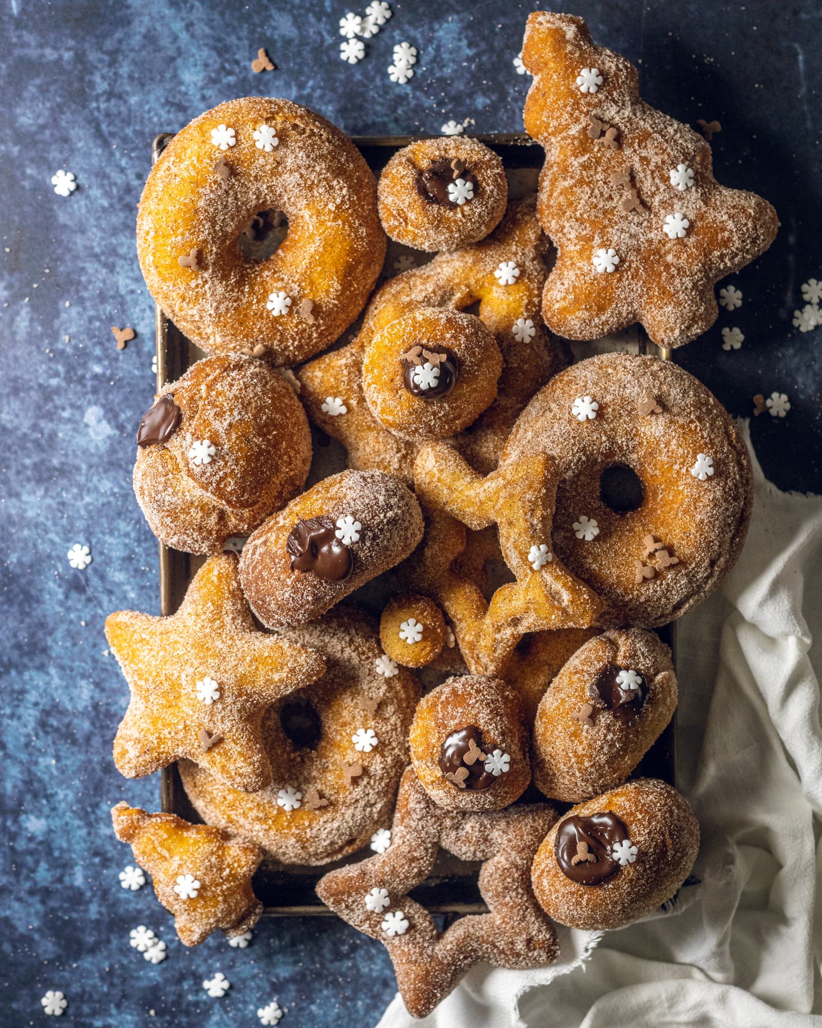 A selection of vegan donuts in different shapes and sizes coated with cinnamon sugar resting on a baking tray.