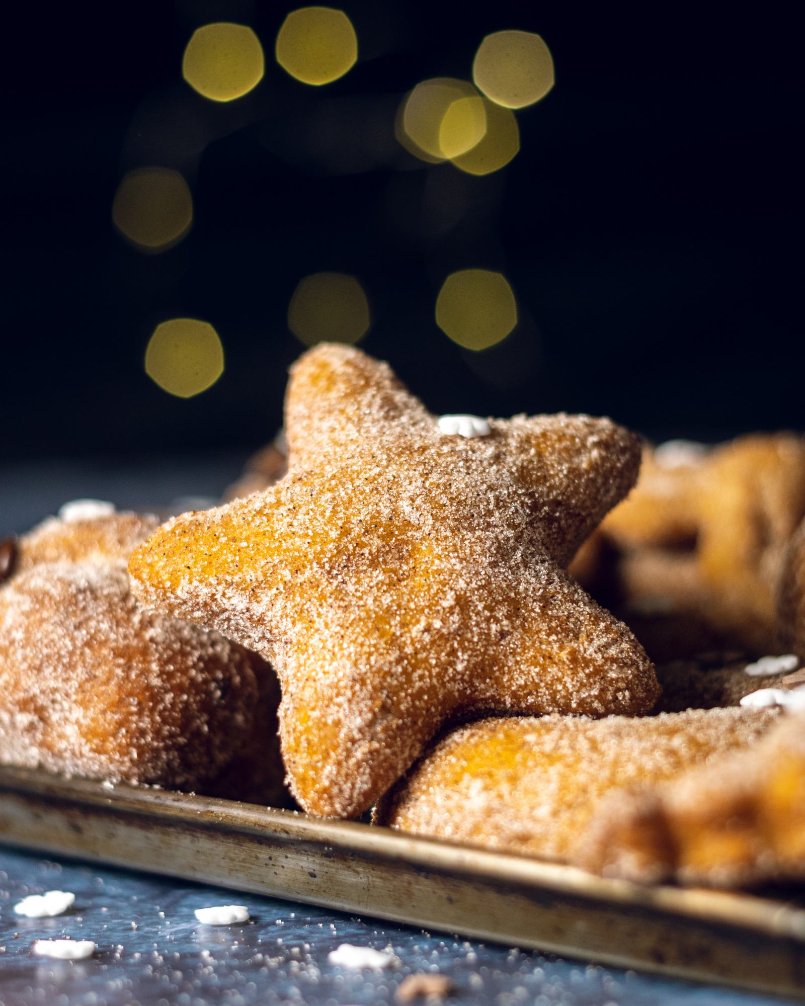 A star shaped vegan cinnamon donut the edge of a baking tray filled with donuts and there are fairy lights visible in the background.