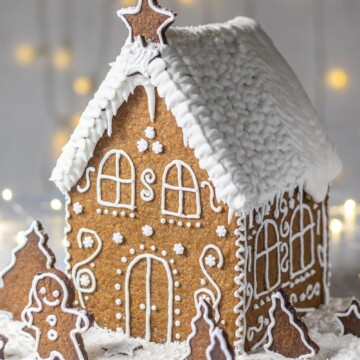 Vegan Gingerbread House, decorated with white buttercream and 'Ho Ho' written on the back, with white roof, photographed on a light backdrop with fairy lights