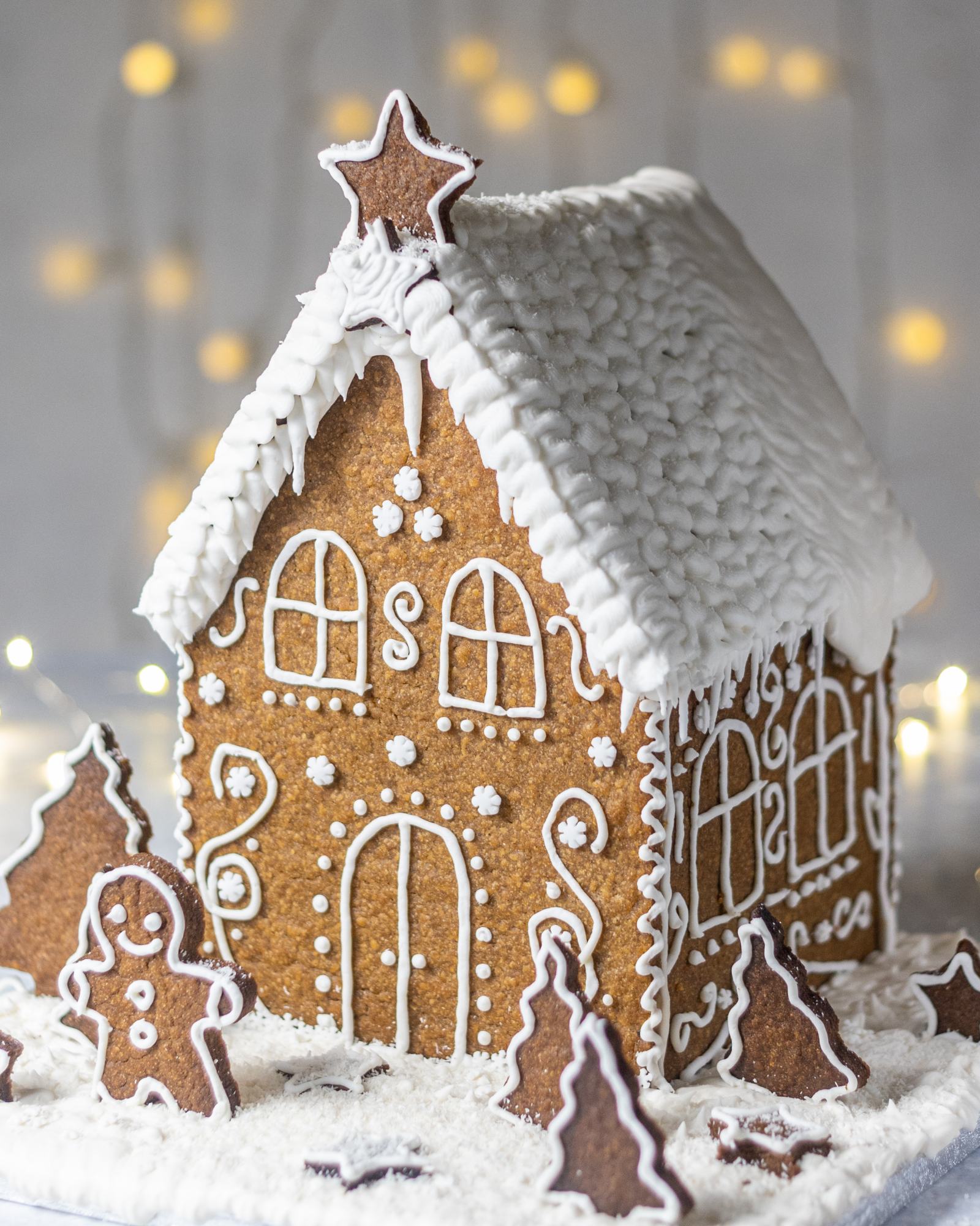 Vegan Gingerbread House, decorated with white buttercream and 'Ho Ho' written on the back, with white roof, photographed on a light backdrop with fairy lights