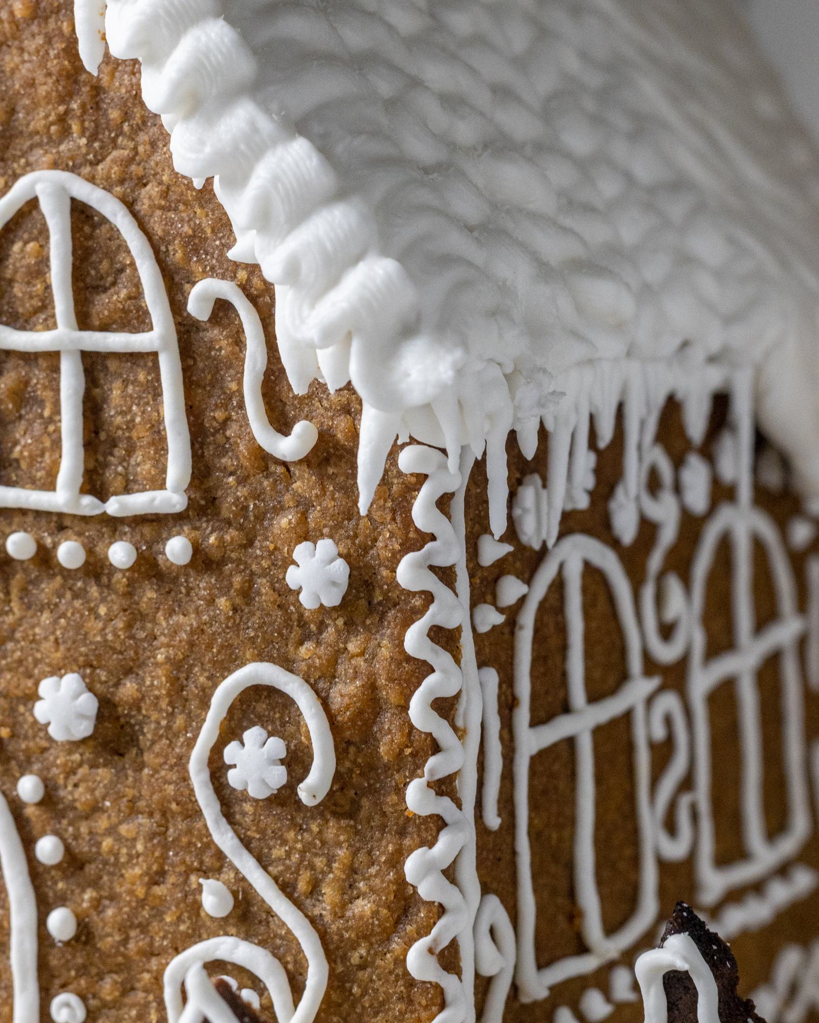 Vegan Gingerbread House, decorated with white buttercream and 'Ho Ho' written on the back, with white roof, photographed on a light backdrop with fairy lights