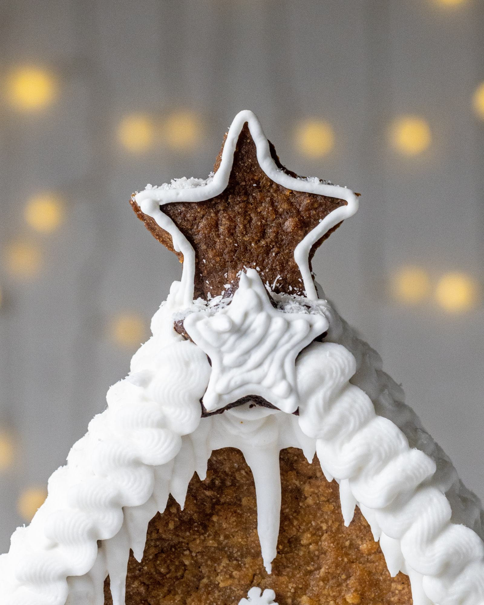 Vegan Gingerbread House, decorated with white buttercream and 'Ho Ho' written on the back, with white roof, photographed on a light backdrop with fairy lights