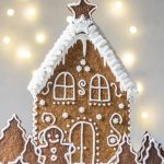 Vegan Gingerbread House, decorated with white buttercream and 'Ho Ho' written on the back, with white roof, photographed on a light backdrop with fairy lights