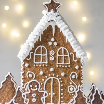 Vegan Gingerbread House, decorated with white buttercream and 'Ho Ho' written on the back, with white roof, photographed on a light backdrop with fairy lights