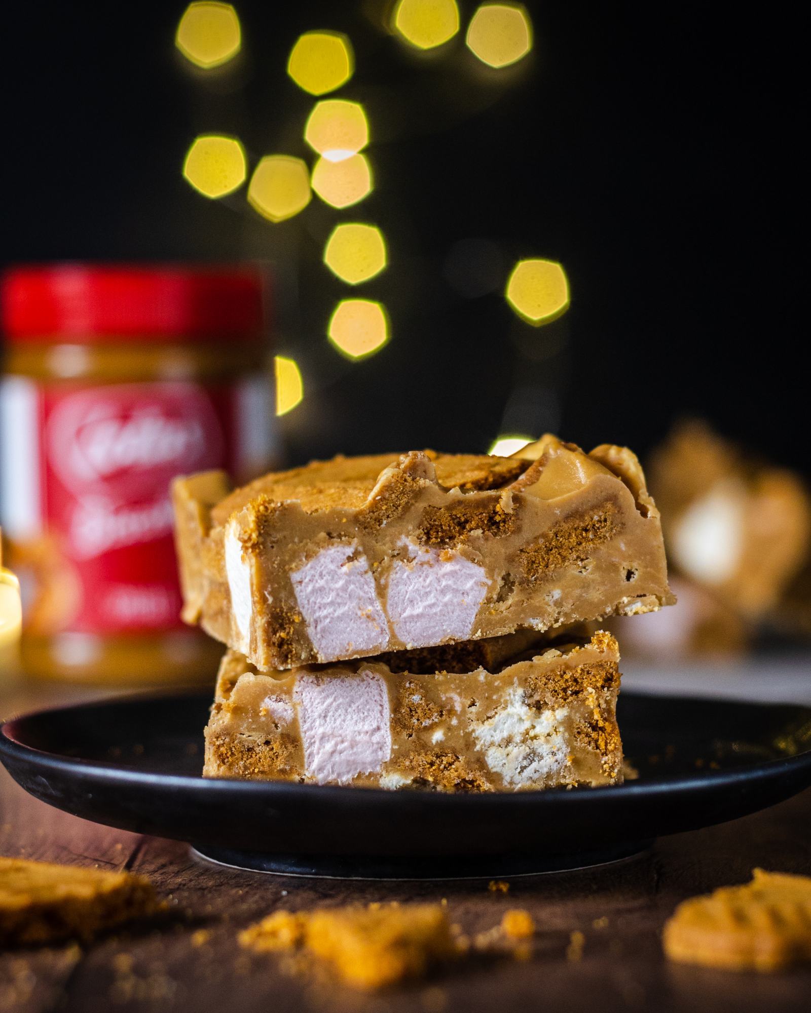 A stack of two vegan biscoff rocky road bars on top of each other, served on a black plate. The crunchy Lotus biscuits and soft marshmallows are clearly visible in the rocky road bar. 