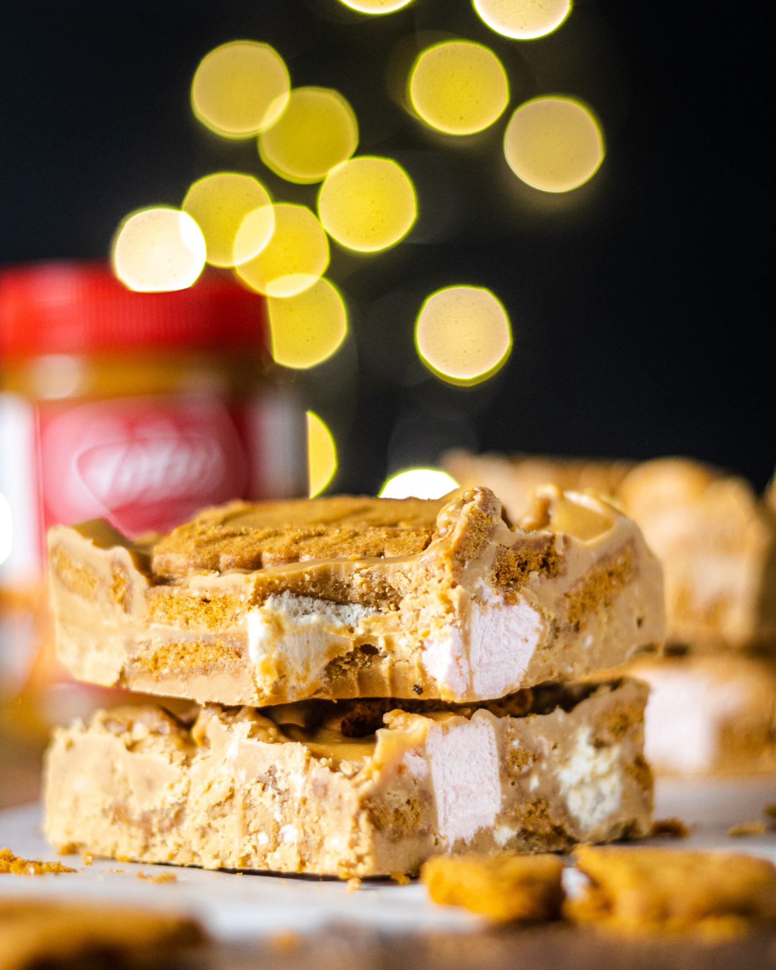 A close up of a stack of Lotus Biscoff Rocky Road bars with a bite taken out of the top one. There are crunchy Lotus Biscoff Biscuits and marshmallows visible and the entire bar has a caramel-like colour. In the background  you can see a blurry biscoff spread jar and fairy-lights in front of a dark background.