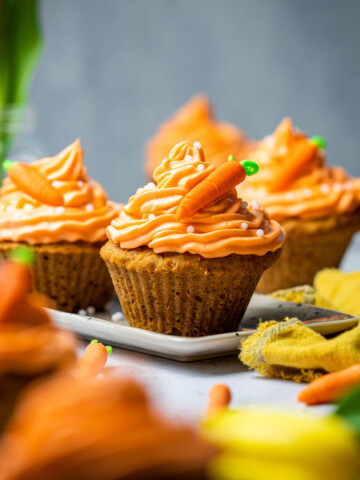 carrot cake cupcakes on a serving platter with mini sugar carrots on top on a table surrounded by flowers