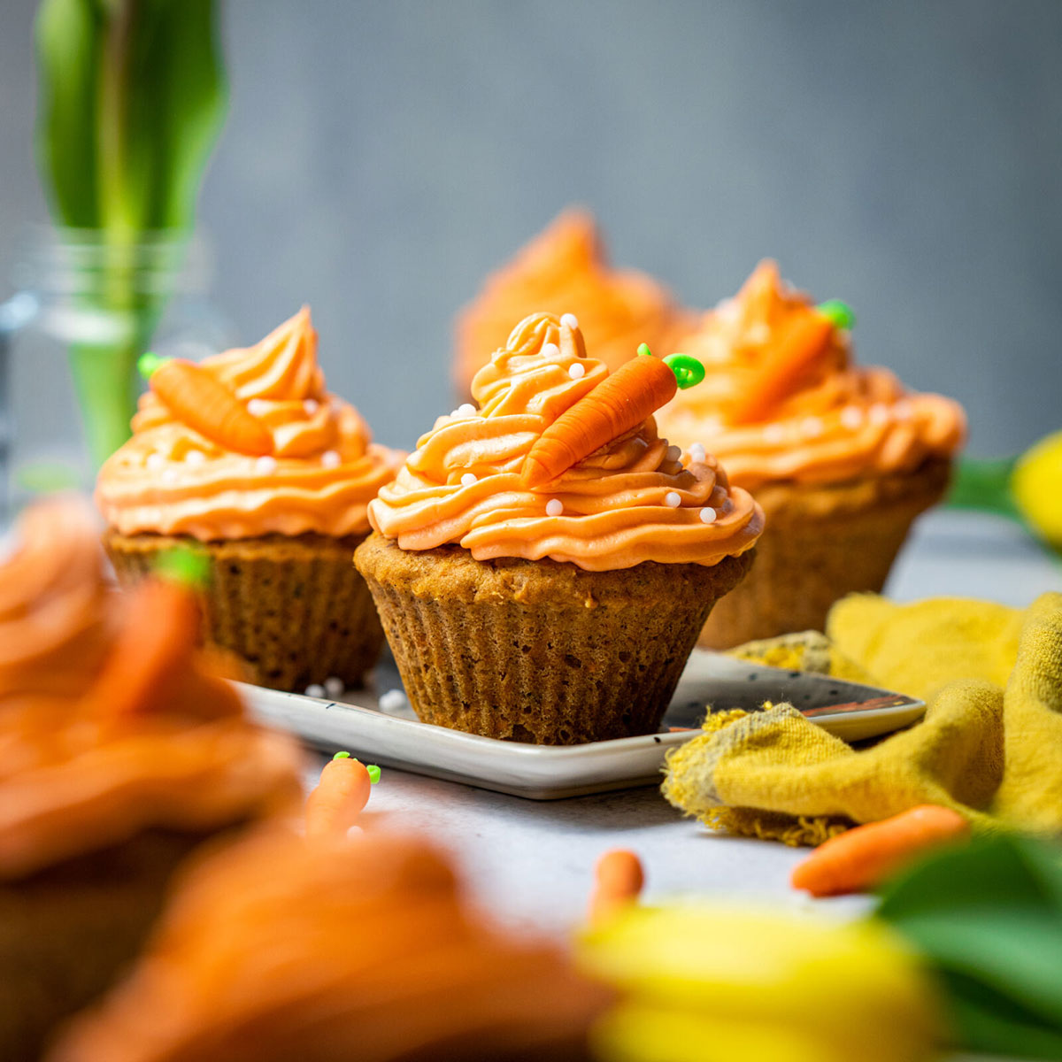 carrot cake cupcakes on a serving platter with mini sugar carrots on top on a table surrounded by flowers