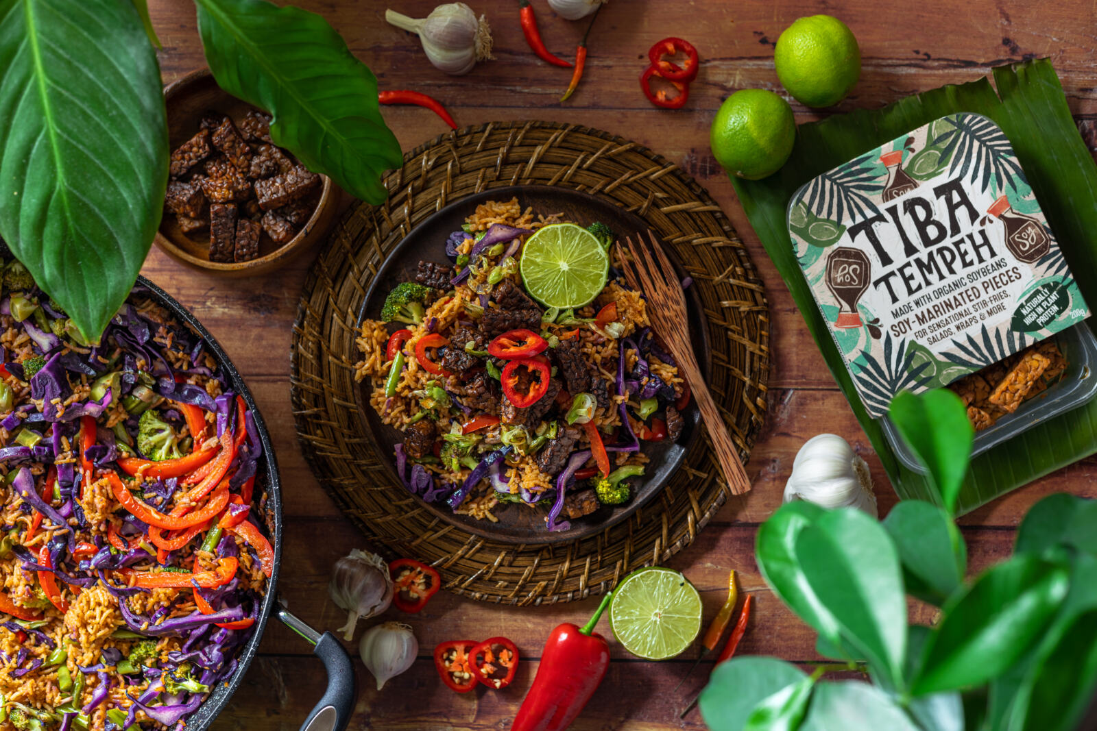 vegan nasi goreng in a bowl on a wooden tabletop next to the tempeh packaging, plants and a wok full of the dish