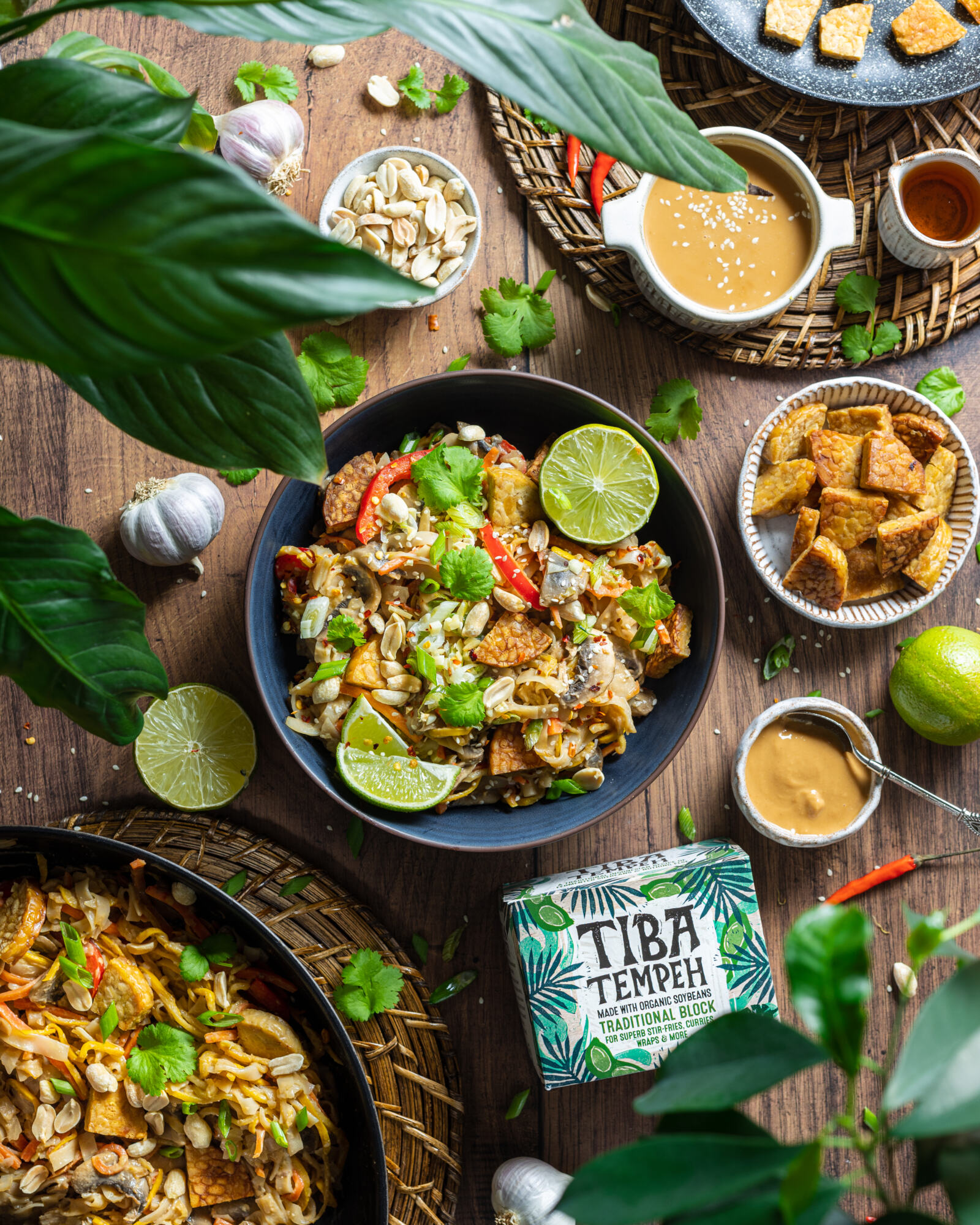 a bowl of vegan pad thai on a wooden table with ingredients and plants surrounding the bowl