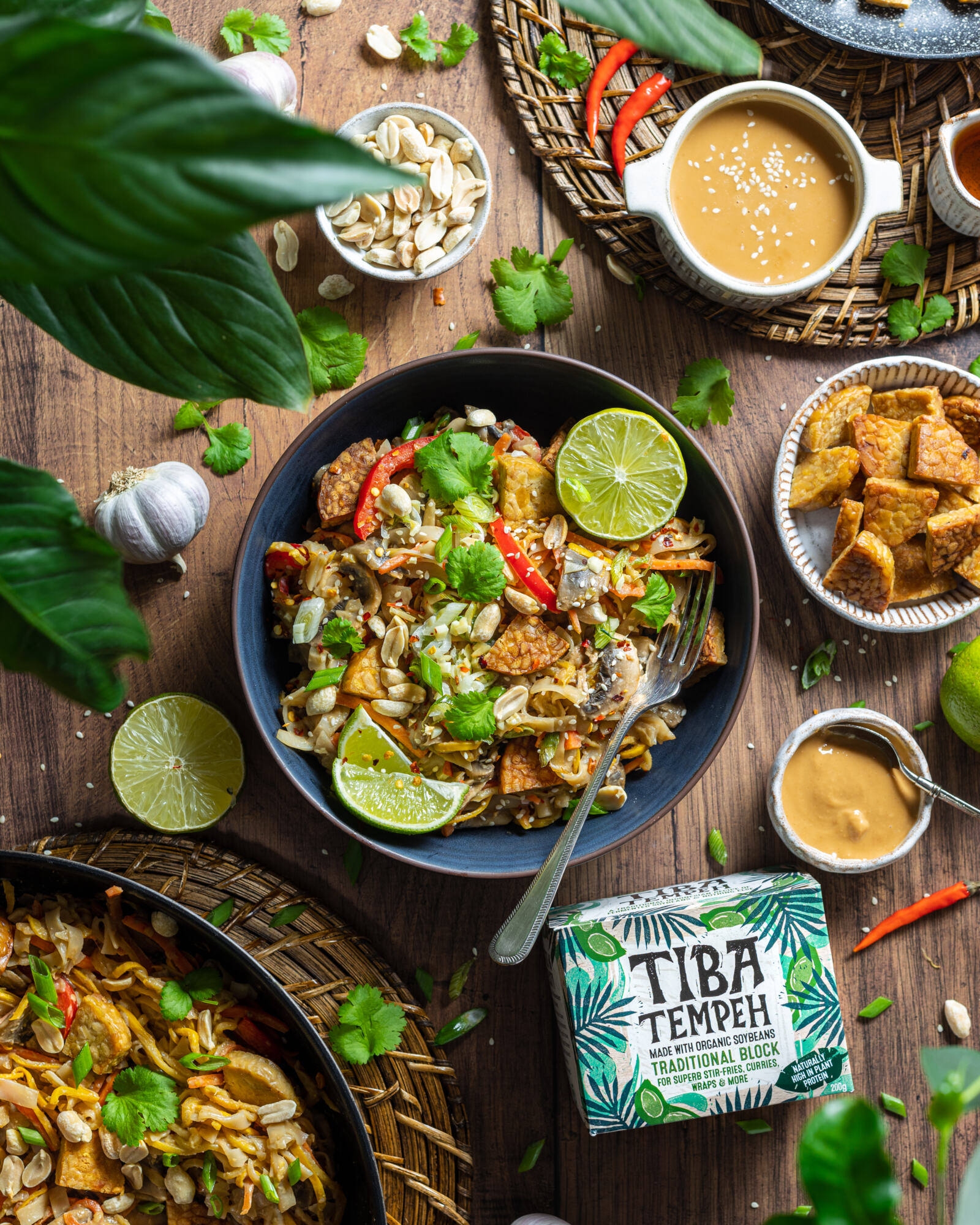 a bowl of vegan pad thai on a wooden table with ingredients and plants surrounding the bowl