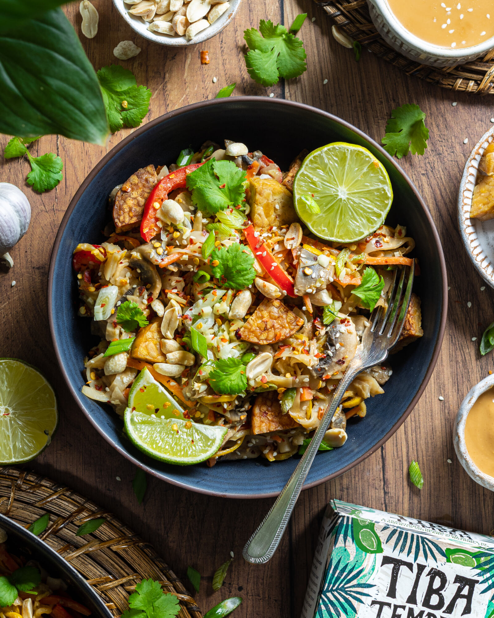 a bowl of vegan pad thai on a wooden table with a fork in the bowl and fresh lime on the side