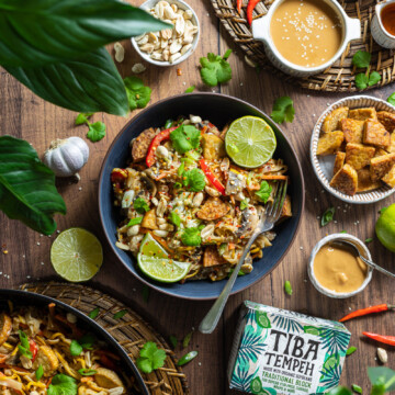 a bowl of vegan pad thai on a wooden table with ingredients and plants surrounding the bowl