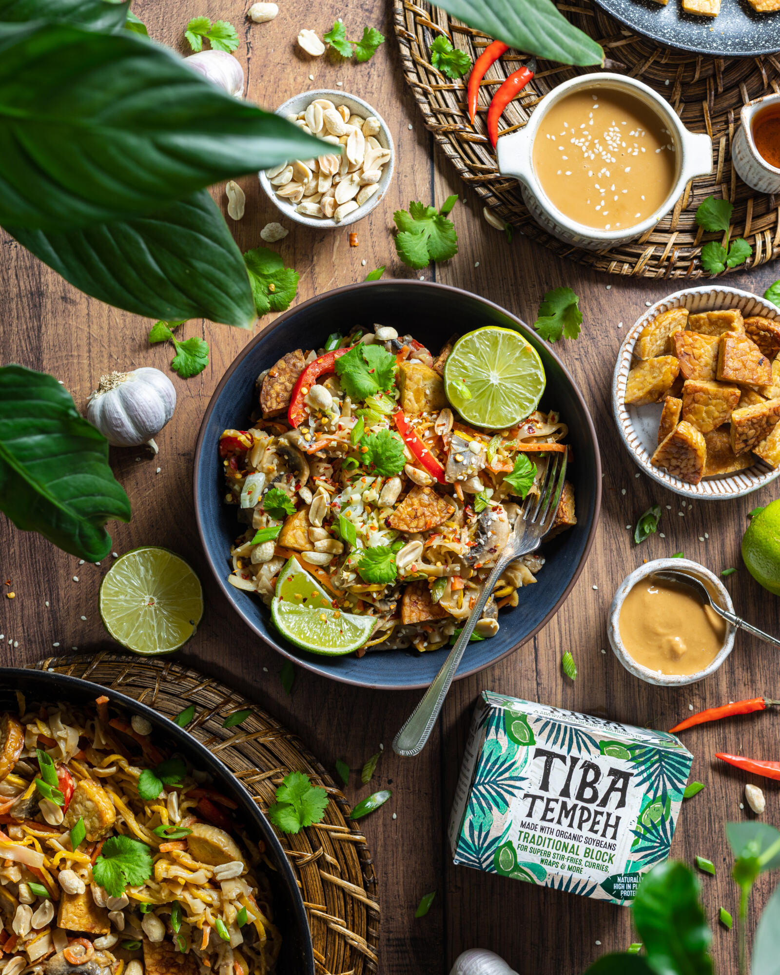 a bowl of vegan pad thai on a wooden table with ingredients and plants surrounding the bowl